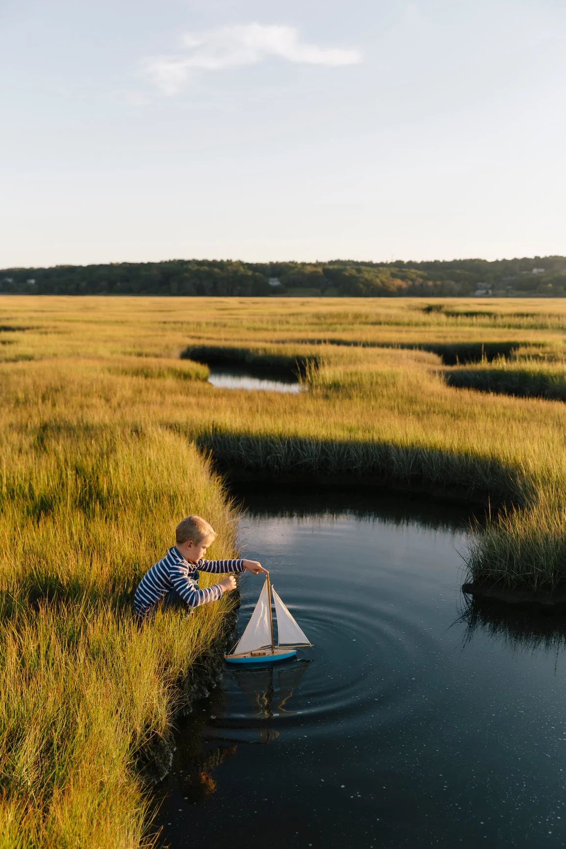 A young boy with blonde hair, wearing a striped long sleeve shirt, is playing with a toy sailboat in a small creek surrounded by tall grass in a marshland at sunset.