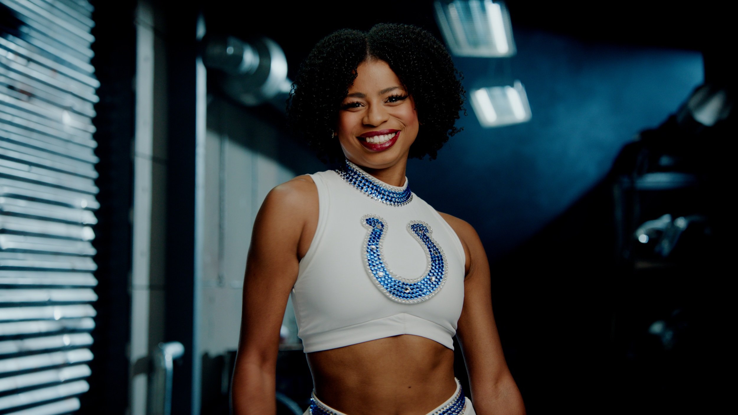 Indianapolis Colts cheerleader wearing a white and blue outfit with a horseshoe emblem, smiling in an indoor setting with metal pipes in the background.