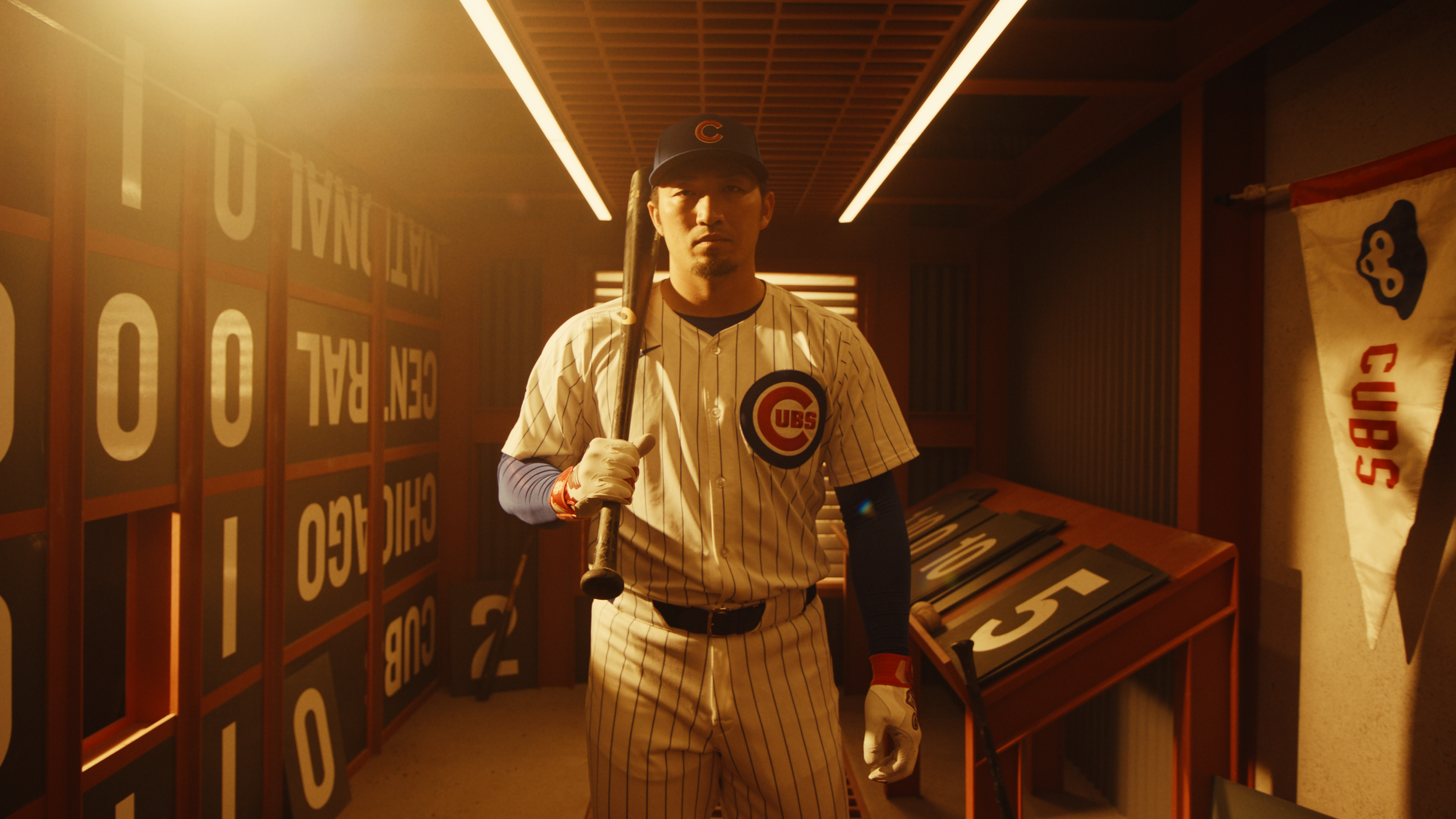 A man in a Chicago Cubs baseball uniform holding a bat, standing in a dimly lit locker room or sports area with team banners and scoreboard signs.