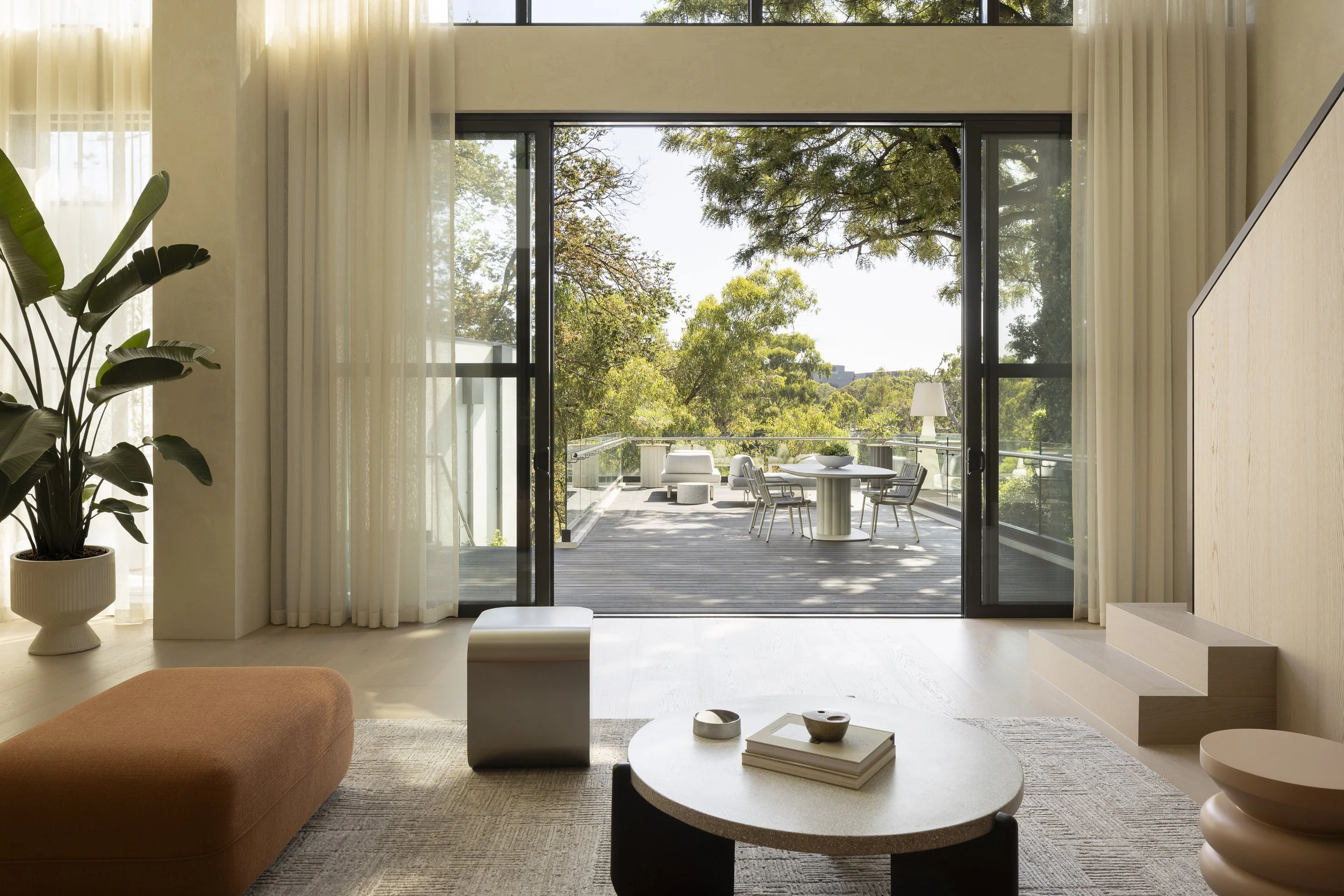 Living room with beige walls, sheer white curtains, a potted plant, and a view of a balcony with an outdoor table, chairs, and trees in the background.
