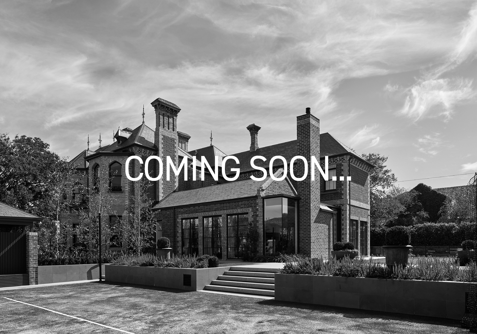 A black-and-white photo of a large brick house with multiple chimneys and tall windows, with trees and a sky with clouds in the background. Overlay text says 'COMING SOON..'.