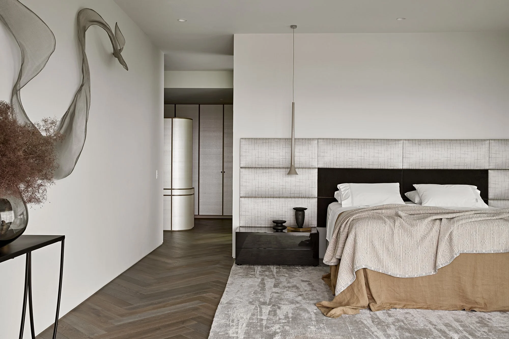 A modern bedroom with a large bed and decorative headboard, wooden flooring, a nightstand with black vases, a tall lamp, wall art, and neutral-colored decor.
