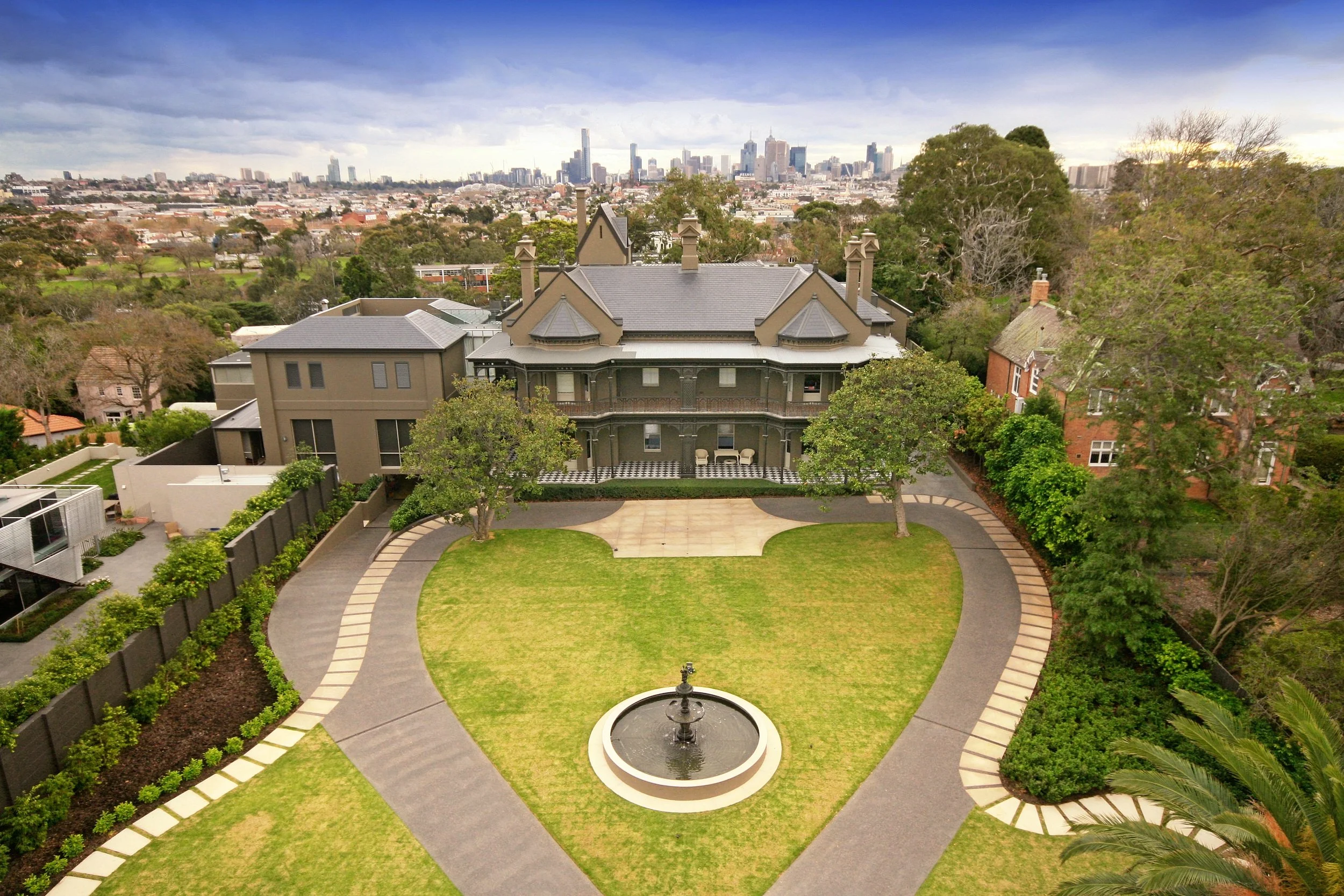 A large, historic mansion with Victorian architectural features, surrounded by trees and a manicured lawn with a central fountain, with the city skyline in the background under a cloudy sky.