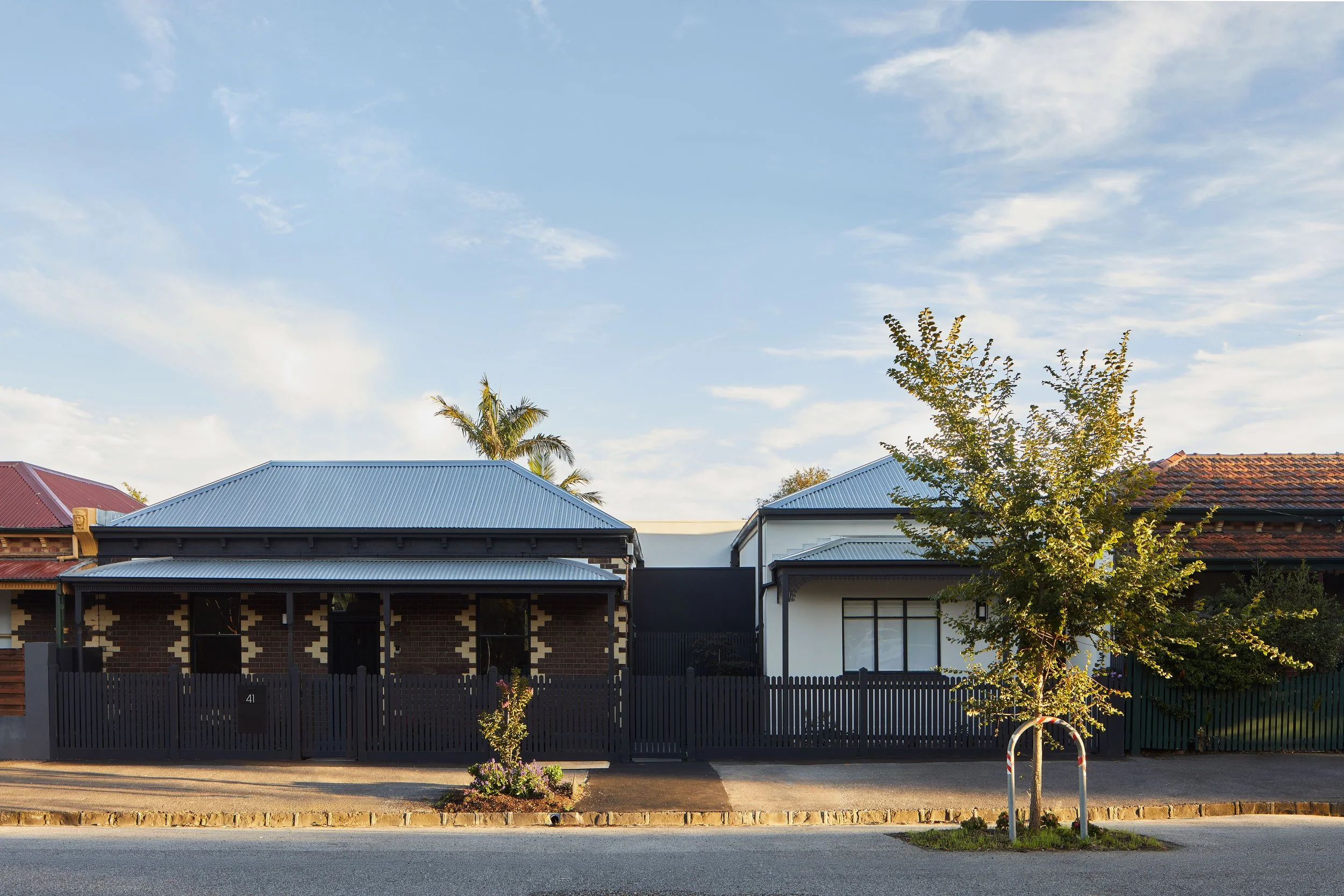 Street view showing houses with metal roofs, a small tree in the foreground, and a clear sky with some clouds.