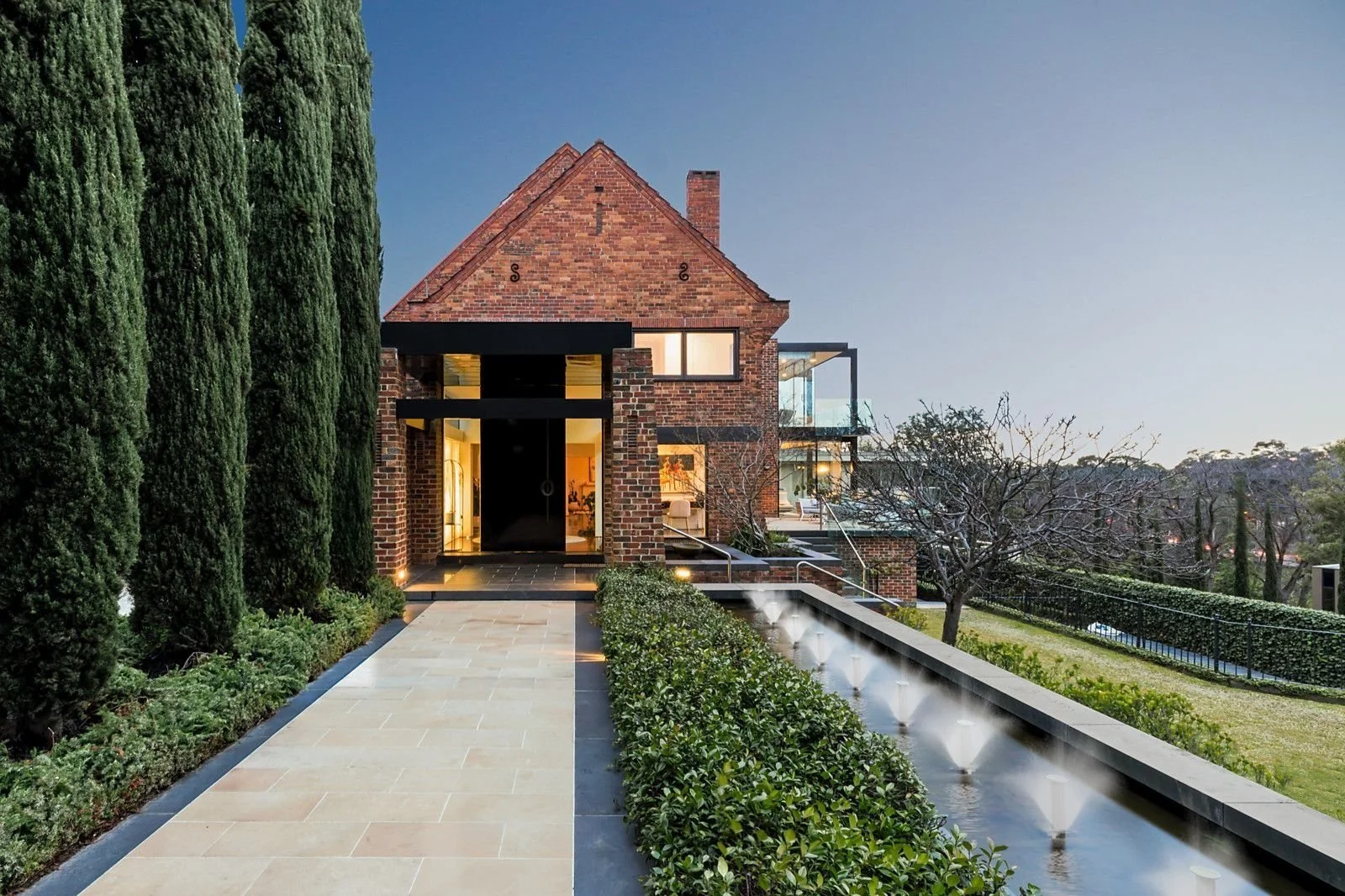 Exterior view of a modern brick house with large windows, glass balcony, and a water feature in the front yard, surrounded by trees and greenery.