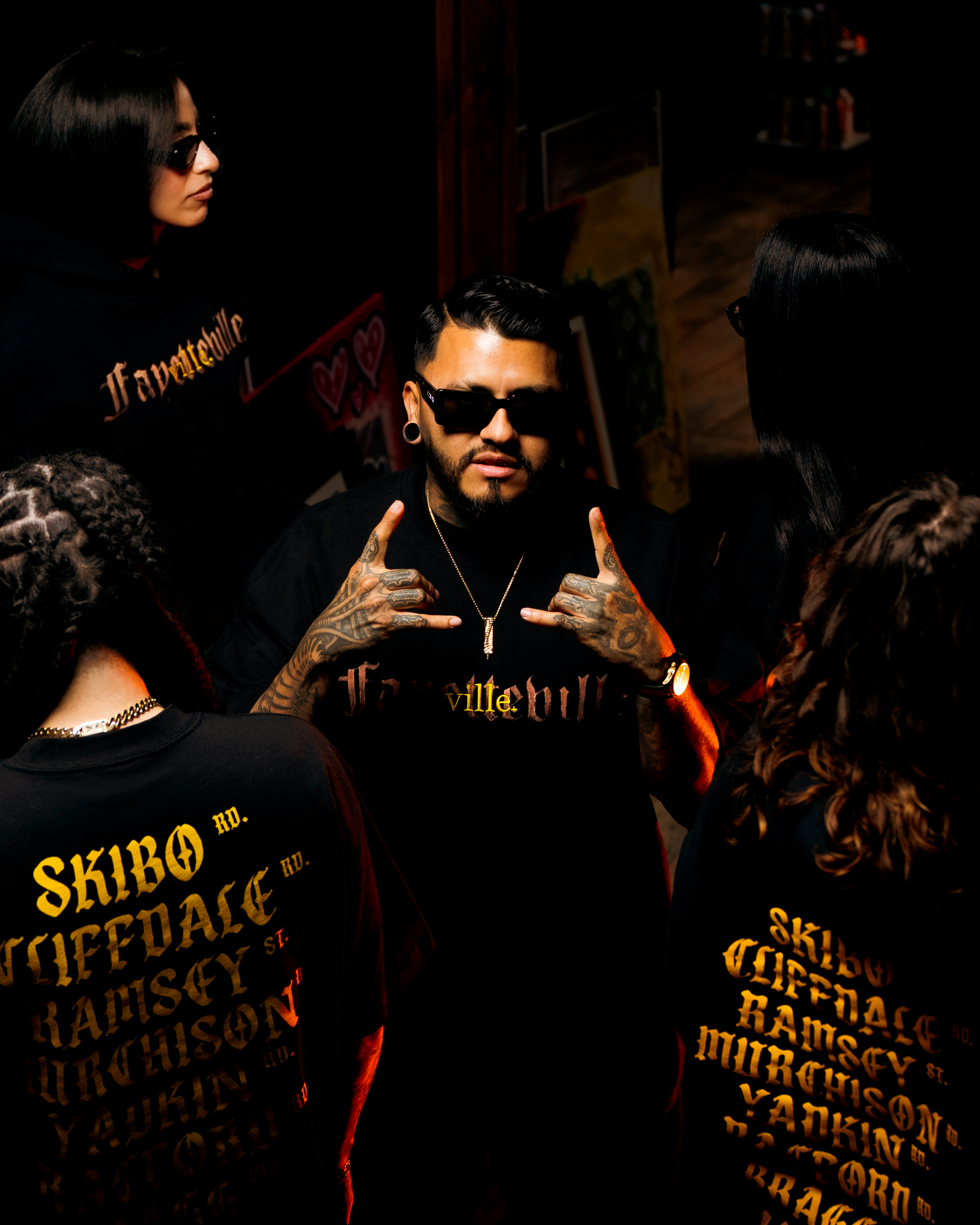 Group of people at an indoor event, with one person in the center making hand gestures, wearing sunglasses, tattoos, and a black shirt with gold text, surrounded by others wearing black clothing and sunglasses.