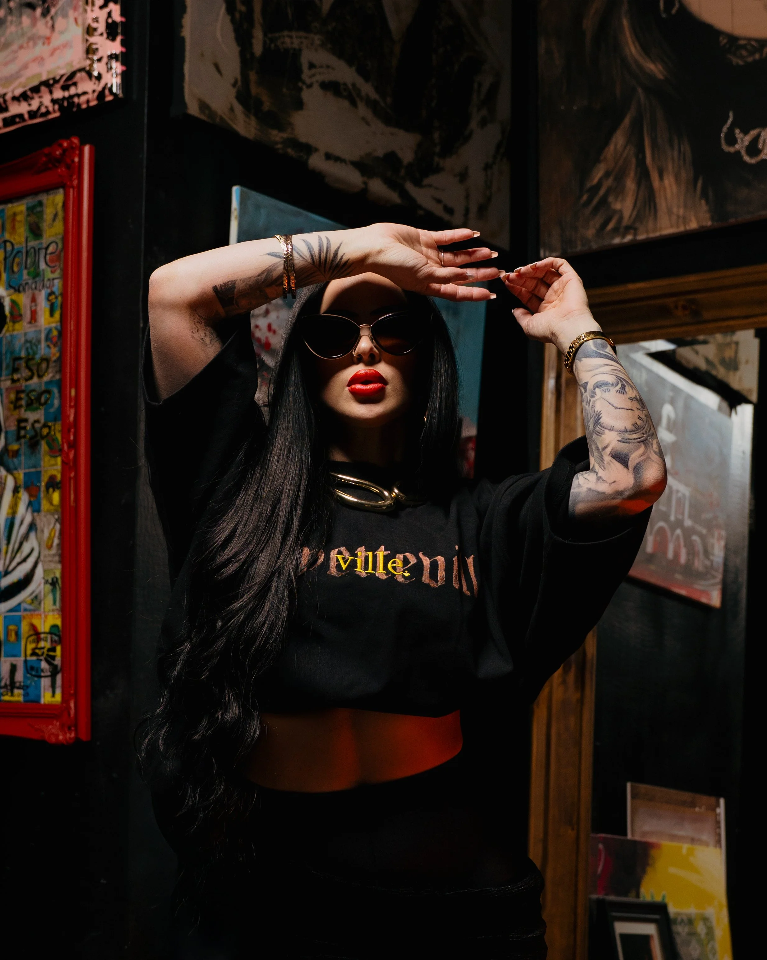 A woman with long black hair, wearing sunglasses, a black t-shirt with the word 'villen' on it, and red lipstick, is standing in front of colorful artwork and posters. She has tattoos on her arms and accessories including a bracelet and a chain neckl