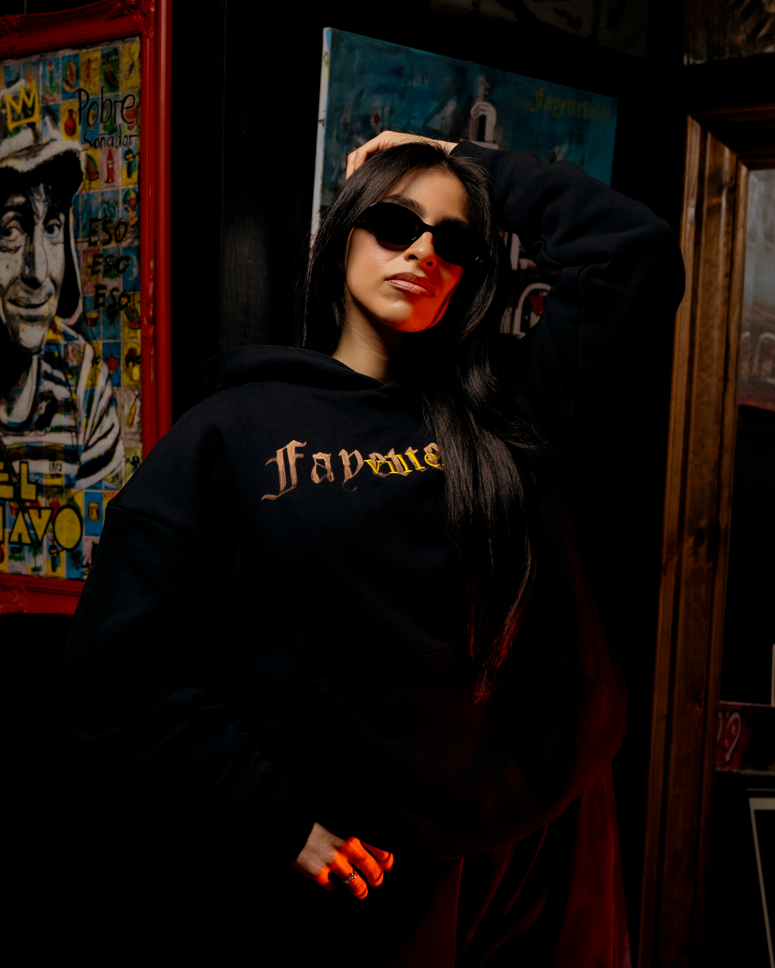 A woman with long dark hair wearing black sunglasses and a black hoodie, posing with her hand on her head, standing in front of colorful posters on a wall in a dimly lit setting.