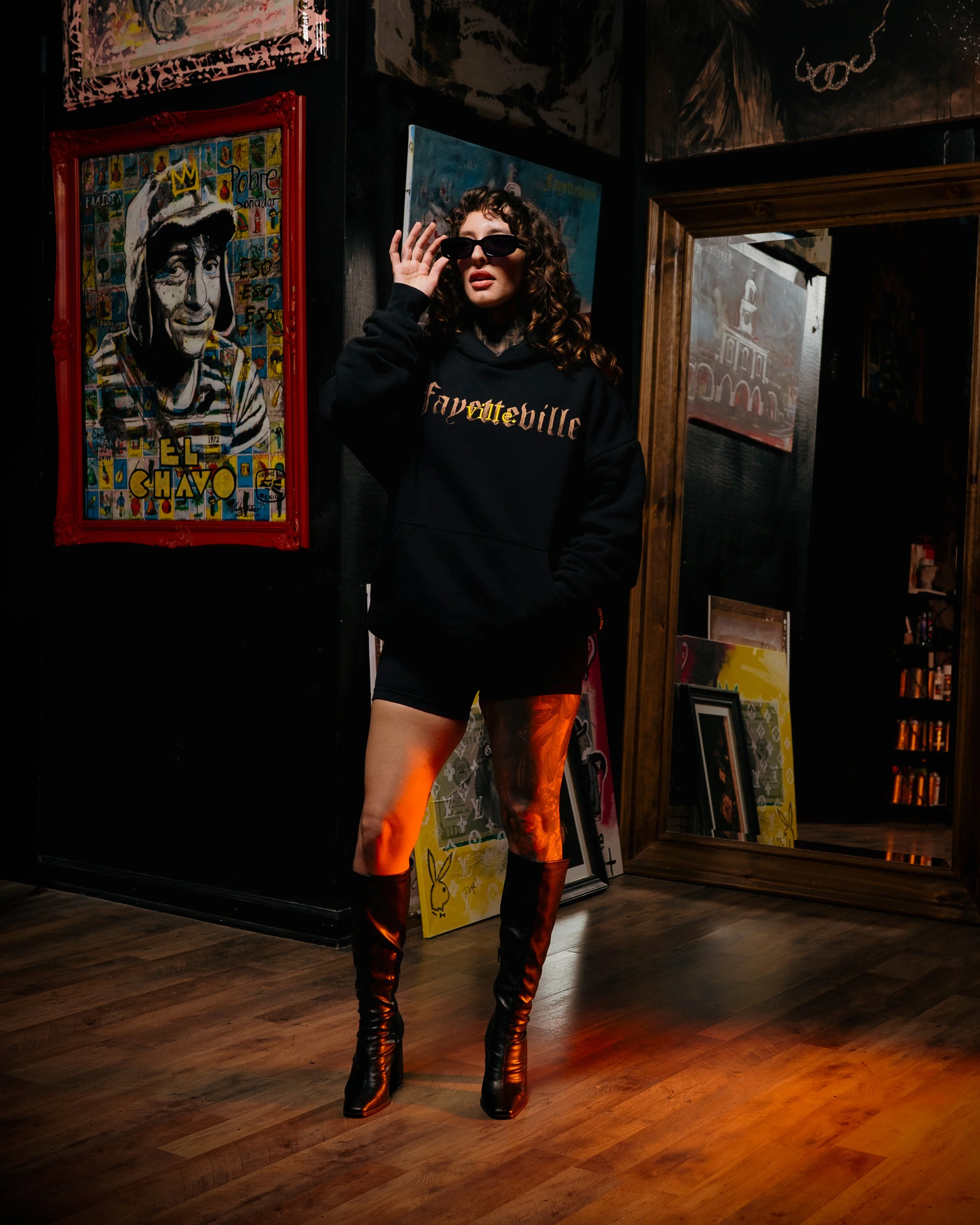 A woman with curly hair and sunglasses wearing a black hoodie with colorful text, black shorts, and tall black boots standing in an art gallery surrounded by colorful artwork and mirrors.