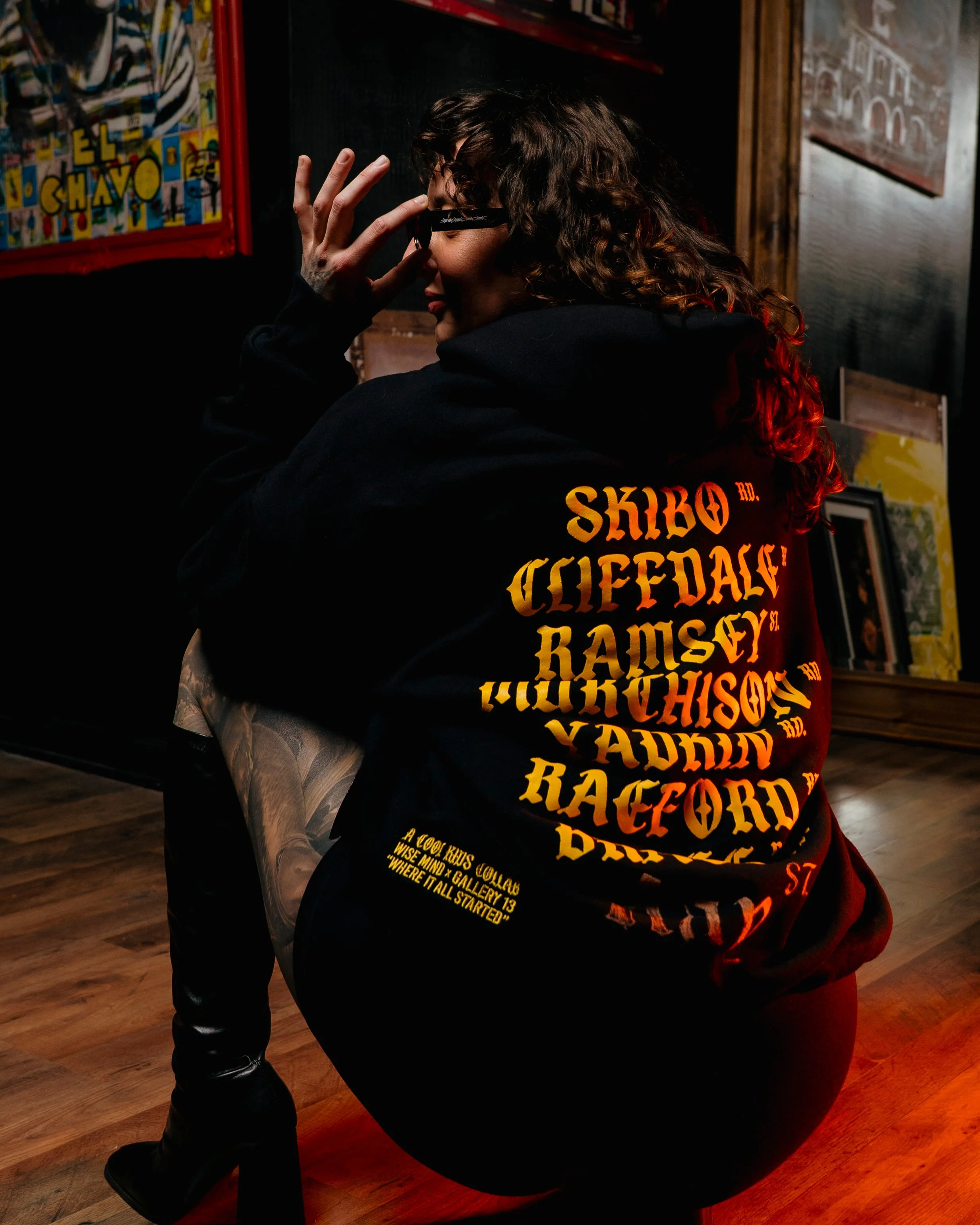 A person with long curly hair and glasses is sitting on a black chair, wearing a black hoodie with orange and yellow text on the back. The person has tattoos on their arm and is slightly leaning forward. They are in a room with wooden floors and artw