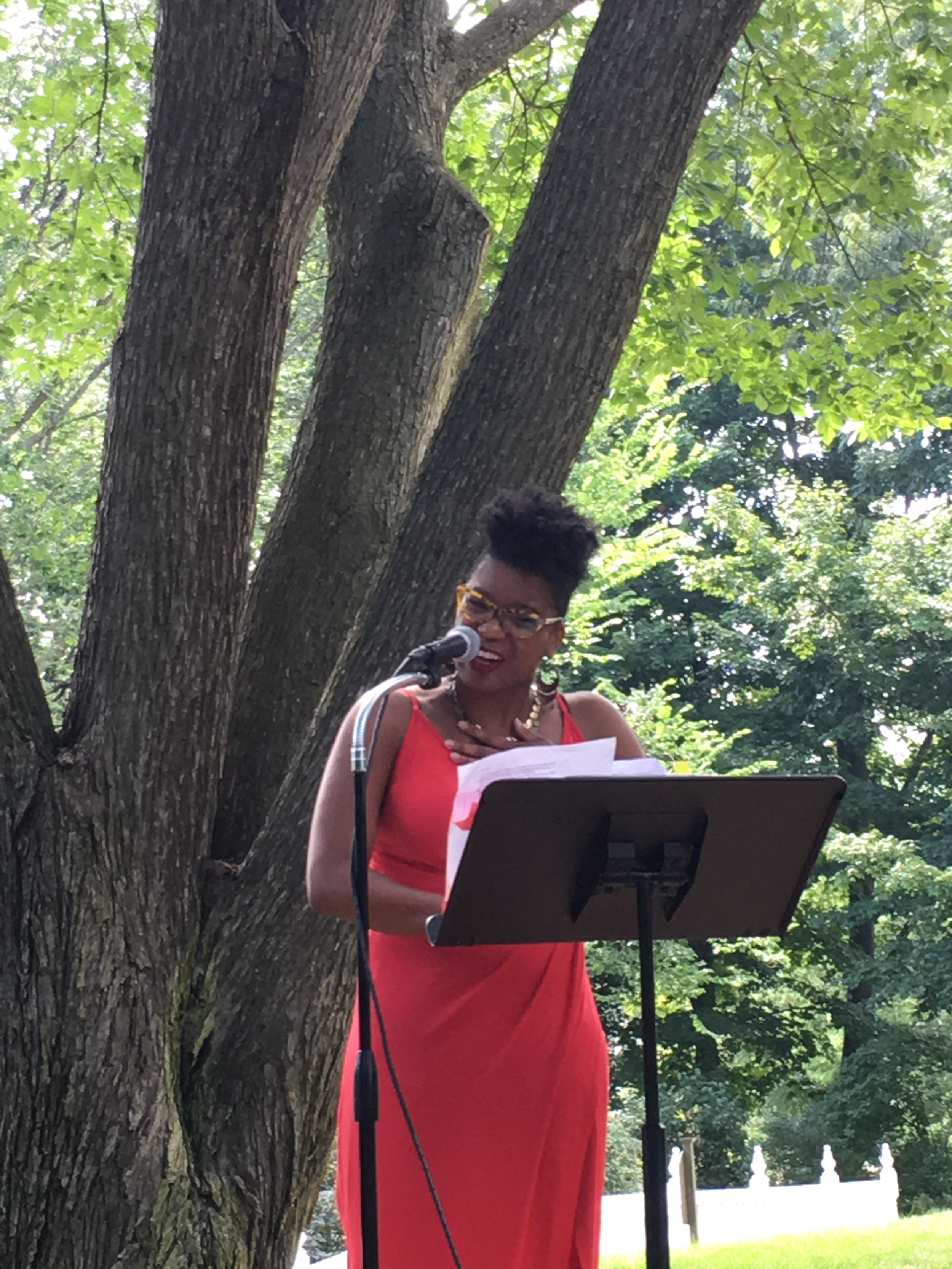 Poet Krysten Hill reading at the New England Poetry Club. She is standing outside, in front of a large tree and behind a music stand, which holds the paper she is reading from. Krysten is a Black woman with short hair, wearing tortoise, round rimmed glasses and chunky earrings, and a long red dress.