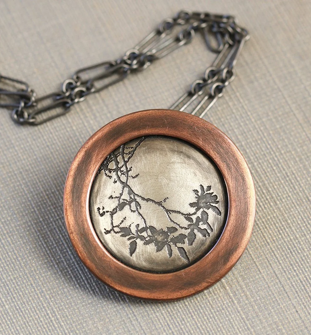 A round silver pendant with a dark patinaed etched tree branch,  and a copper ring border, with a dark silver chain, also made by Dana.