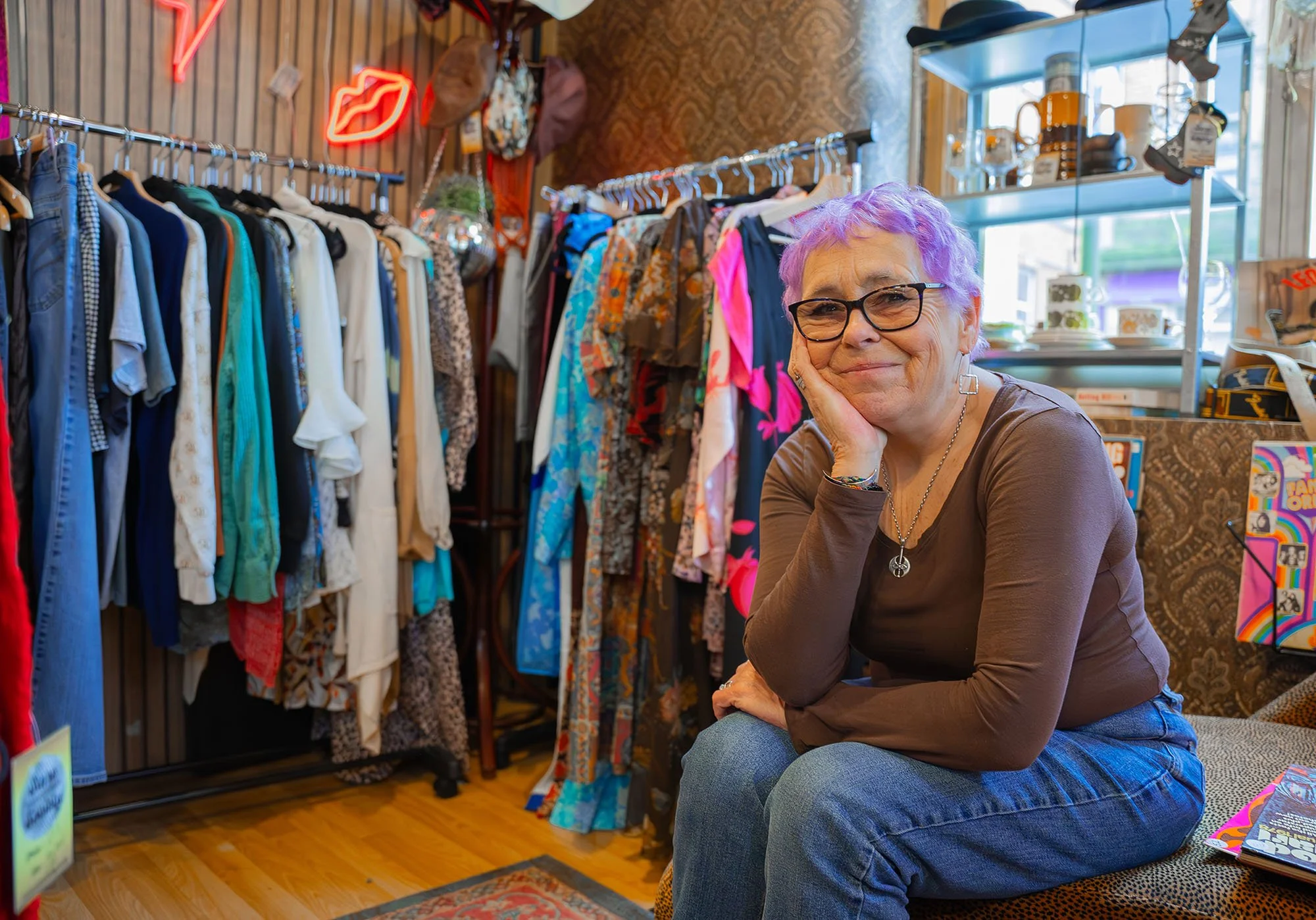 Light skinned woman with short purple hair, dark rimmed glasses, wearing brown top and jeans. She is seated in a thrift shop, in front of a a clothing rack and a window with dishes.