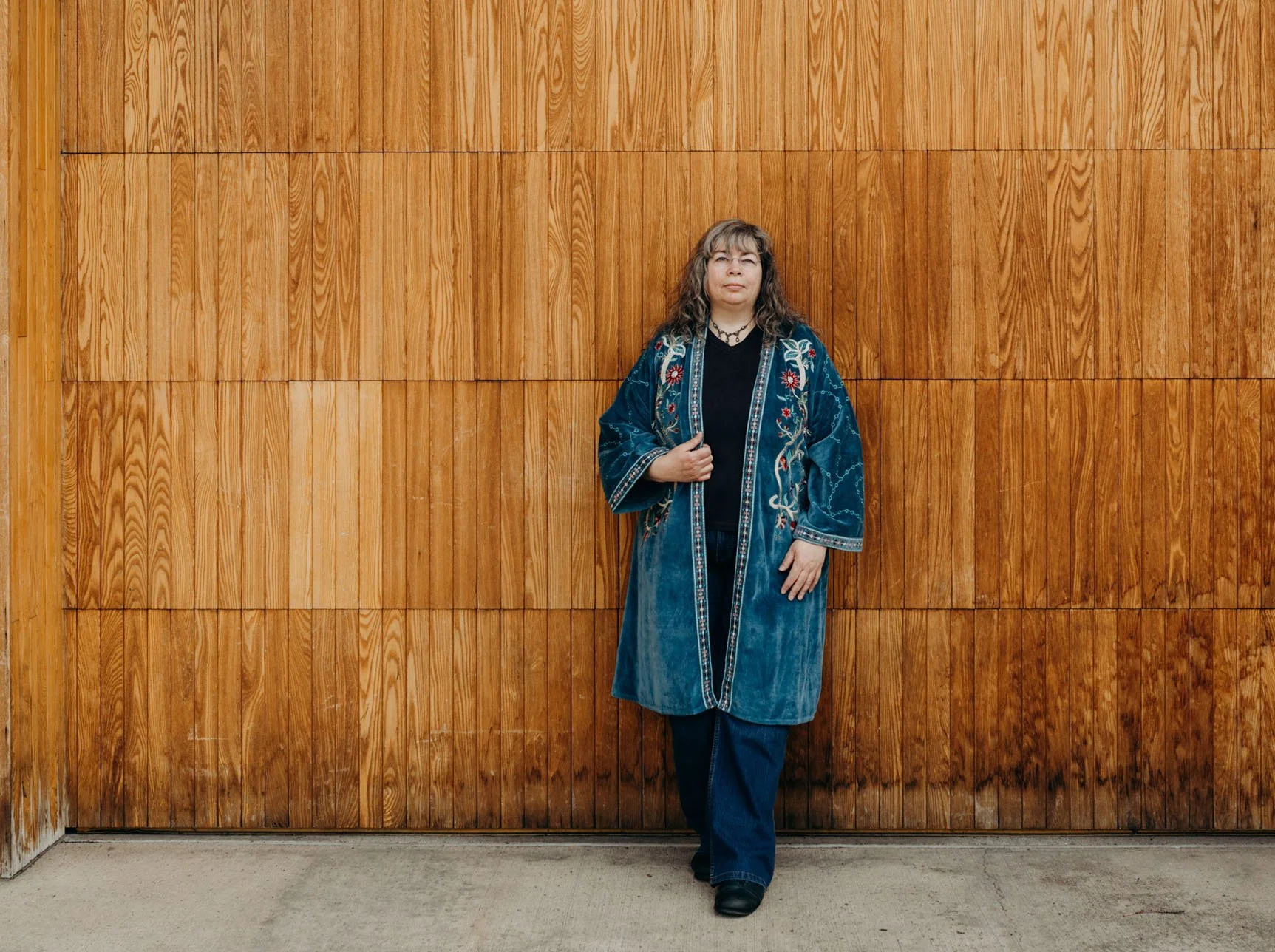 Web designer, Dana Reed, a light skinned woman with long dark hair, wearing long teal velvet coat with floral embroidery, standing in front of wood backdrop