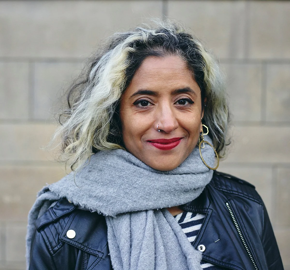 Medium skinned woman with medium length grey and black hair wearing a scarf, large hoop earrings and leather jacket, in front of a brick wall.