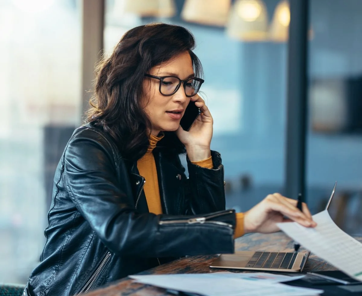 Medium skinned woman with shoulder length dark hair, wearing black framed glasses and a black leather jacket. She is holding a phone to her ear while holding a pen and paper in the other hand, sitting with a laptop, in front of a large window.