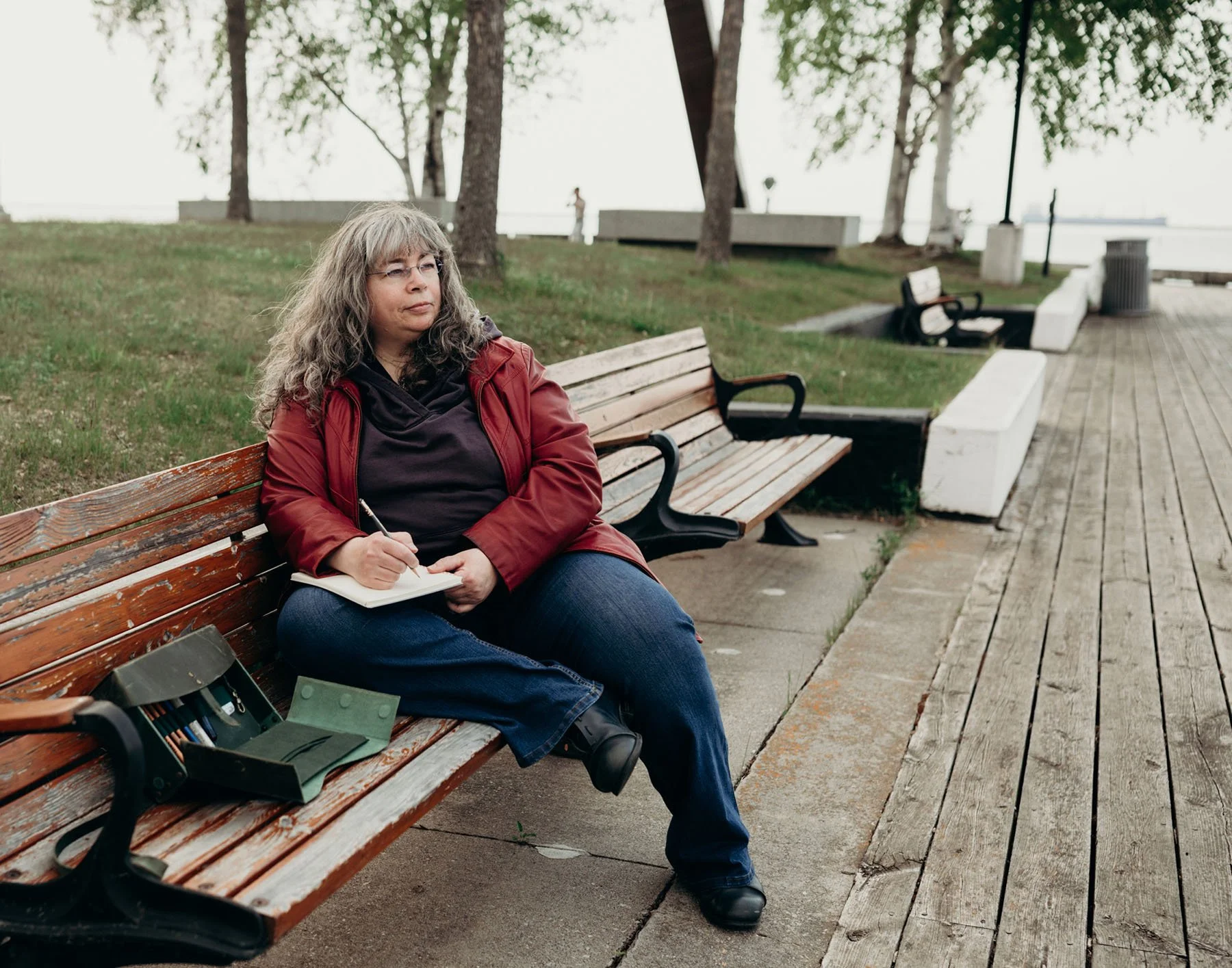 Dana, sitting on a bench, trees in the background, one knee up. She is writing in a notebook, while looking to the right. She is wearing a red leather jacket and glasses.