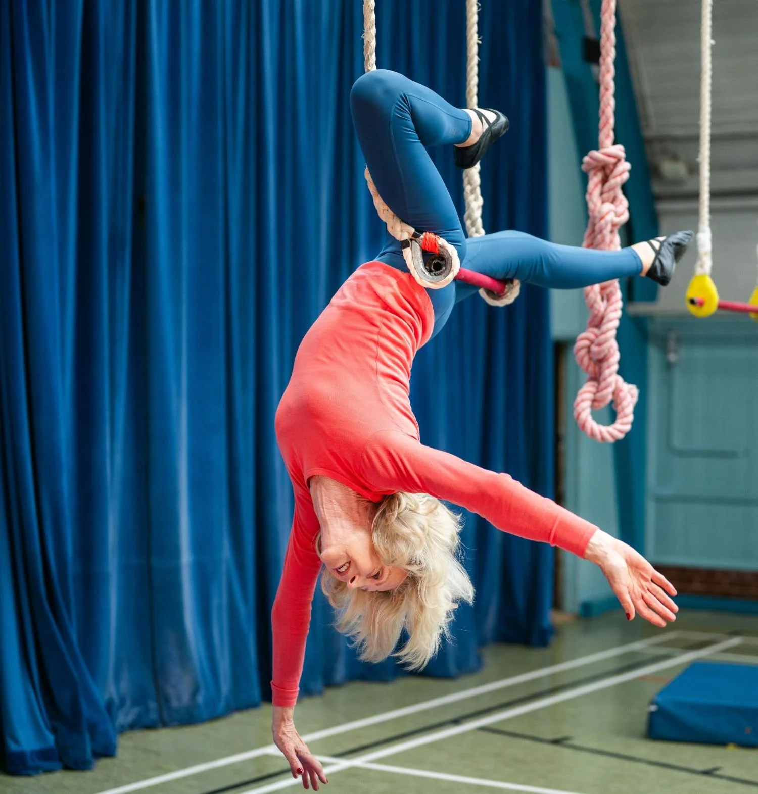 Light skinned older woman with blond hair, in colourful clothes, hanging upside down from a trapeze bar.