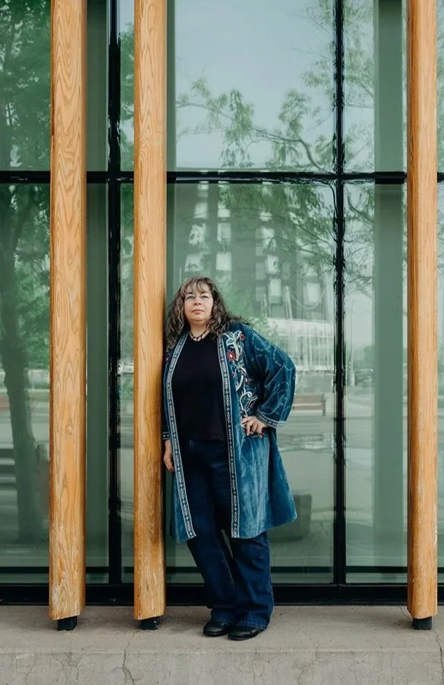 Dana, still wearing her embroidered long teal velvet coat , is leaning on wooden poles, outside, in front of windows with reflections.