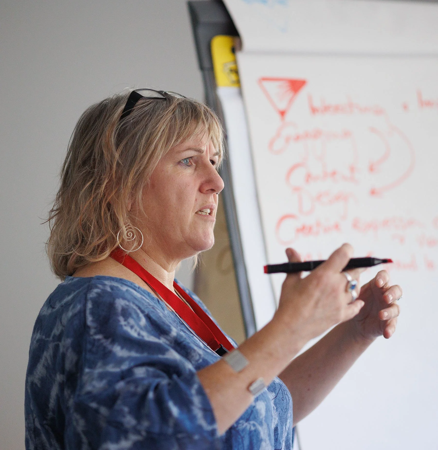 Light skinned woman with medium length blond hair, standing in front of white board, hand in the air, giving a talk.