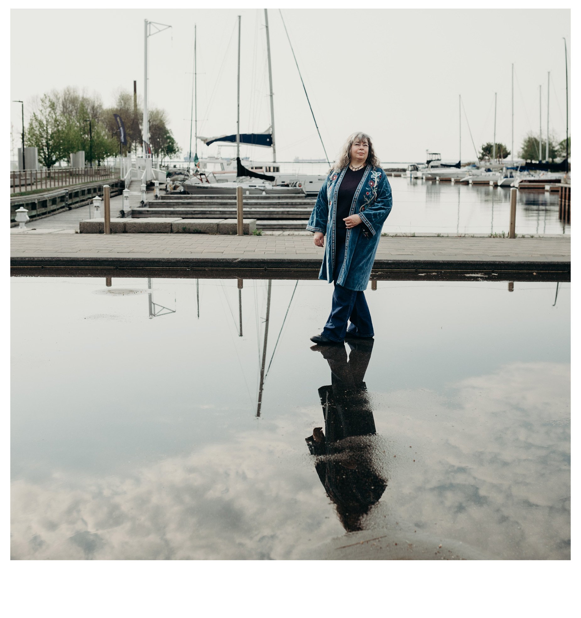 Dana Reed, web designer, standing in shallow water, with her image and clouds reflected in it. She is light skinned, with long grey and dark wavy hair, wearing a teal velvet long coat with floral embroidery. There are sailboats docked behind her.