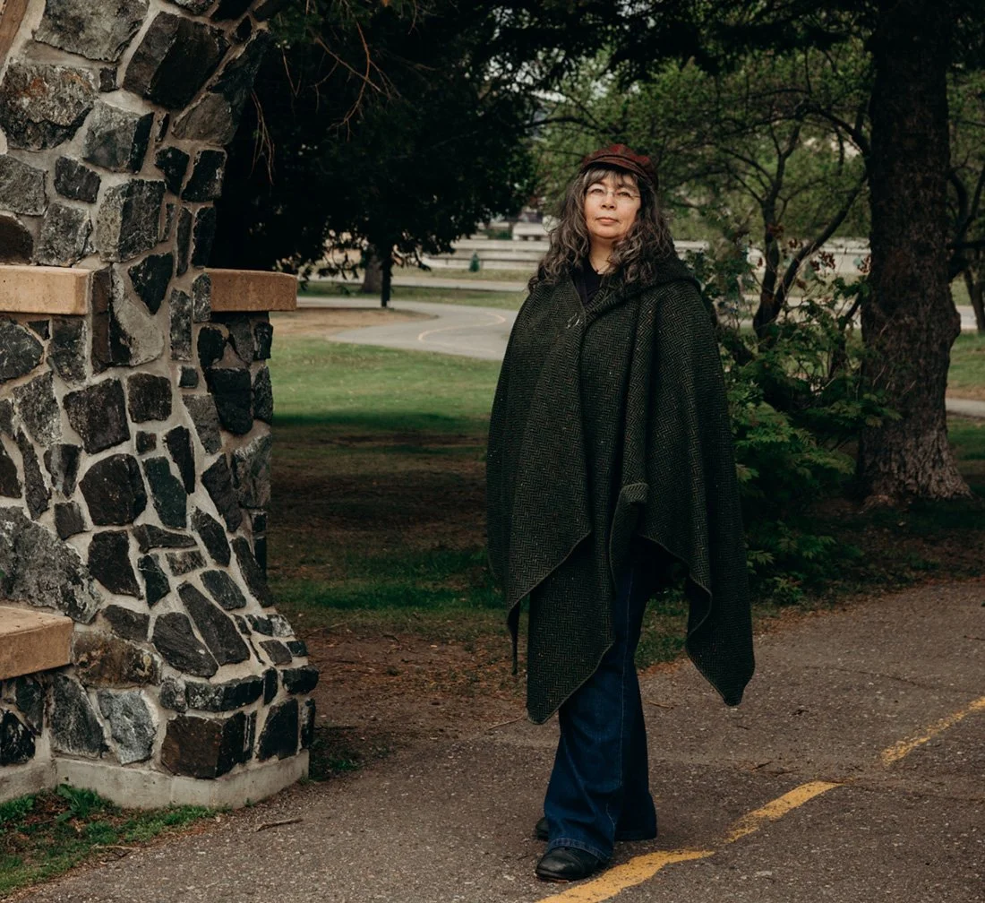 Dana, standing in a stone arch at the Thunder Bay marina with trees in the background, wearing a green tweed cloak, glasses and fabric hat.