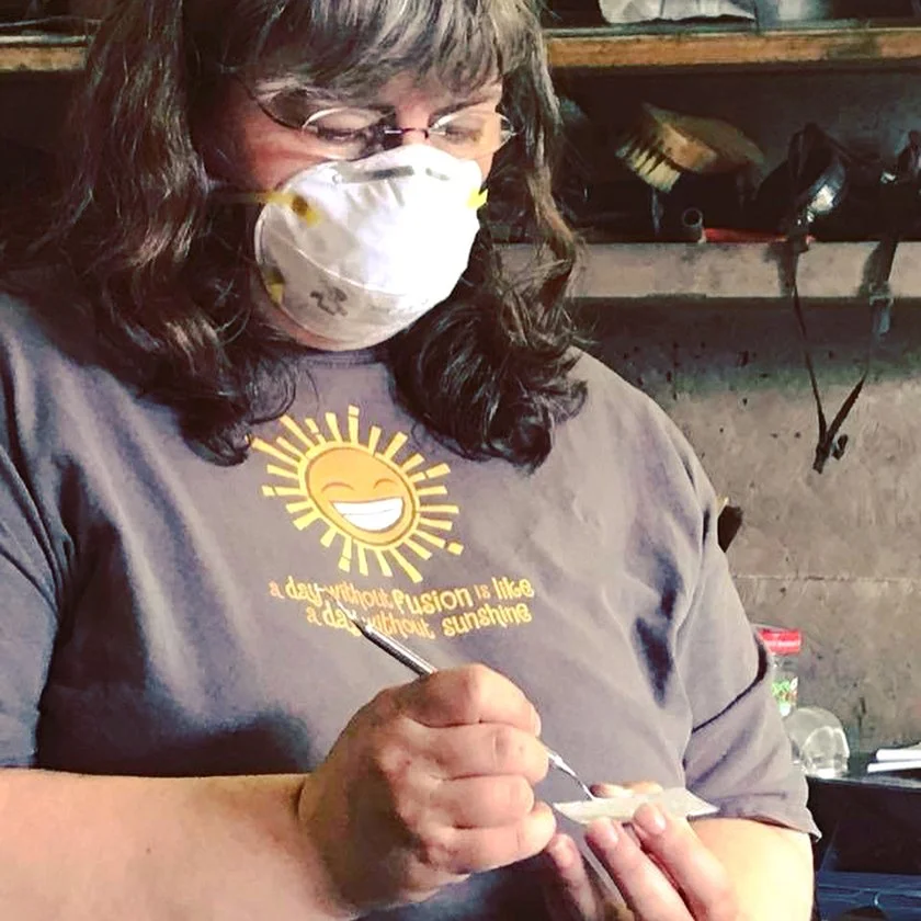 Dana is wearing a dust mask, glasses, and a t-shirt with a sun image and the words a day without fusion is like a day without sunshine. She is painting enamel on a small square of steel.