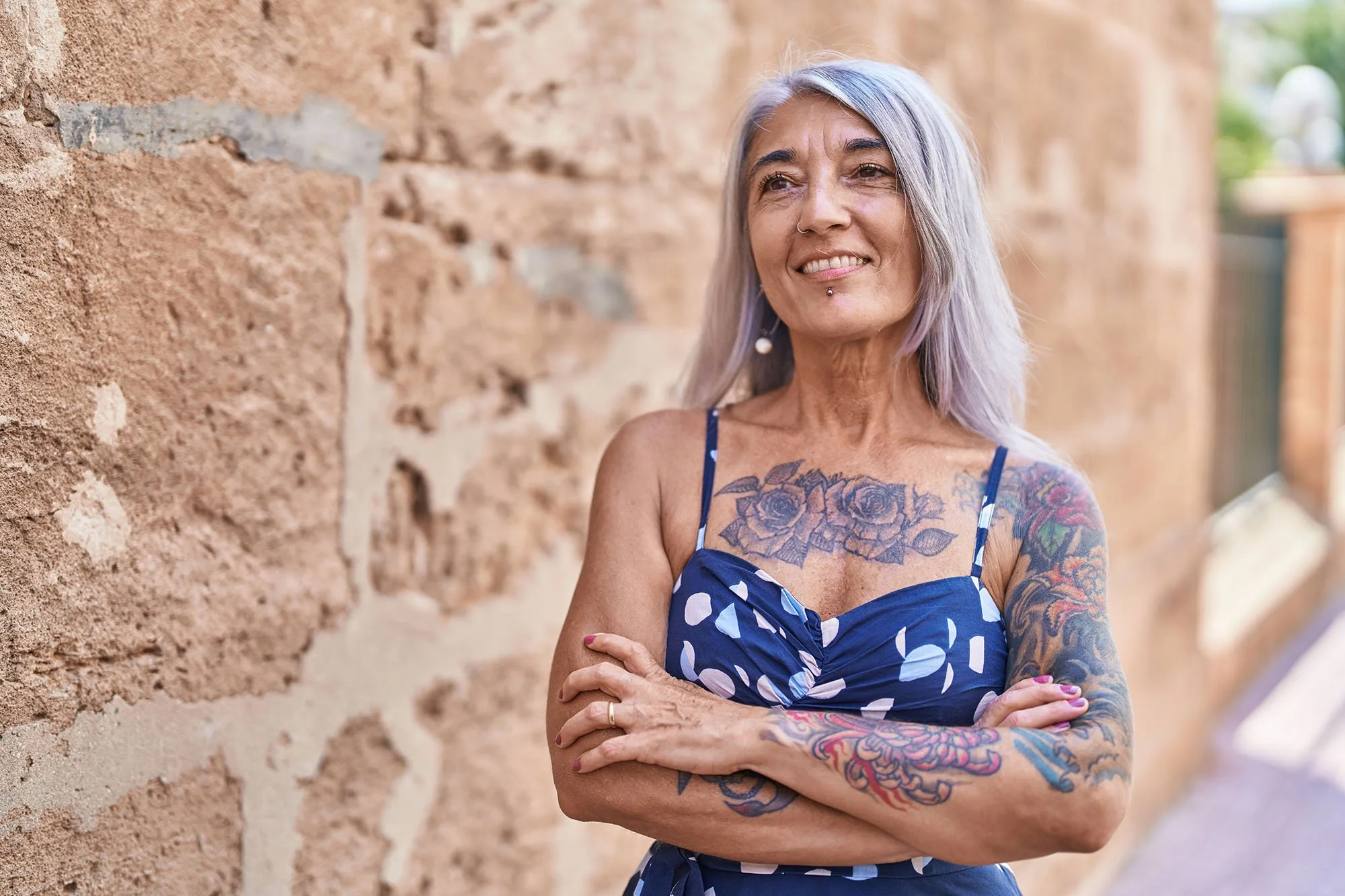 Light-medium skinned woman with long grey hair, tattoos, her arms crossed, wearing a blue sun-dress with dots, standing beside stone wall.