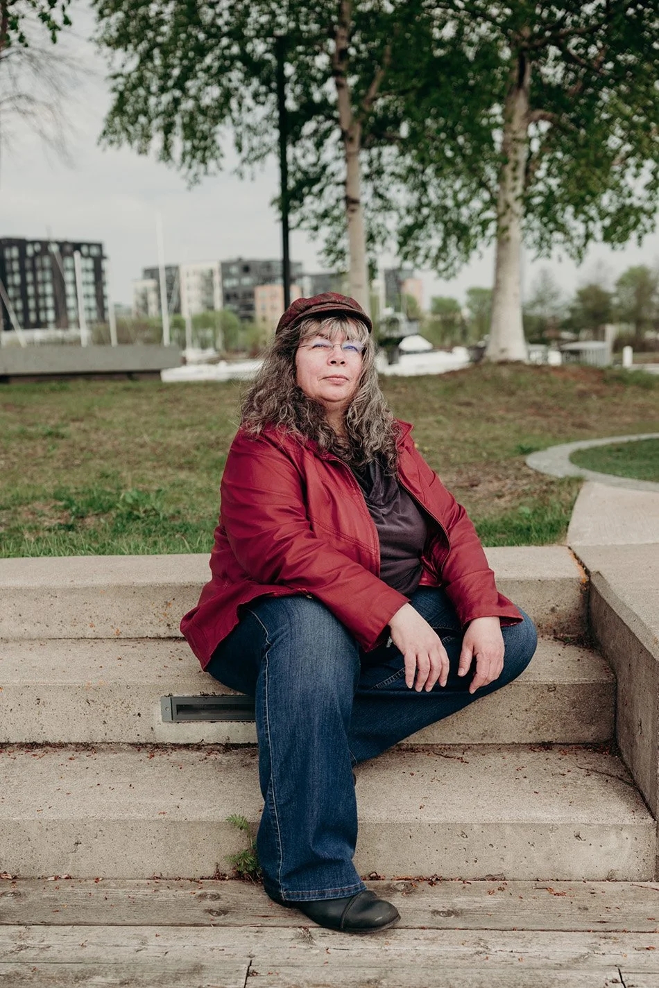 Dana Reed, web designer, sitting on concrete steps in front of birch trees and buildings in the background. She is a light skinned woman with long grey and dark curly hair, wearing a fabric cap, glasses and a red leather jacket.