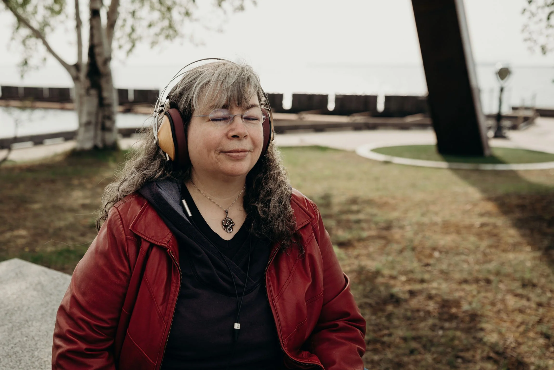 Dana Reed, web designer, a light skinned woman with long grey and dark hair, glasses. She is wearing a read leather jacket and large headphones. Her eyes are closed and she is smiling while sitting in a park.