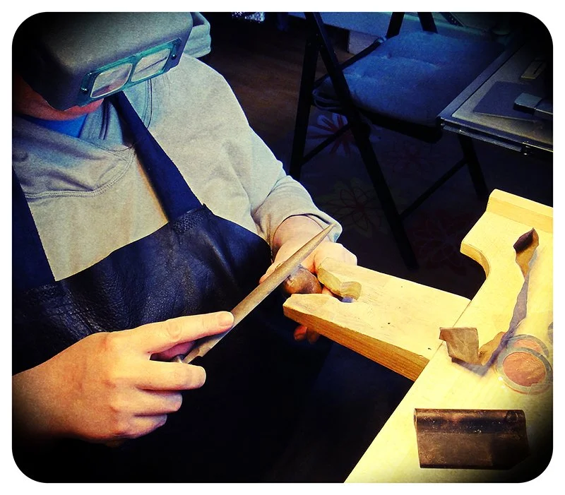 Front angled view of Dana Reed filing metal at a jewellery bench, wearing an apron and magnifying visor.