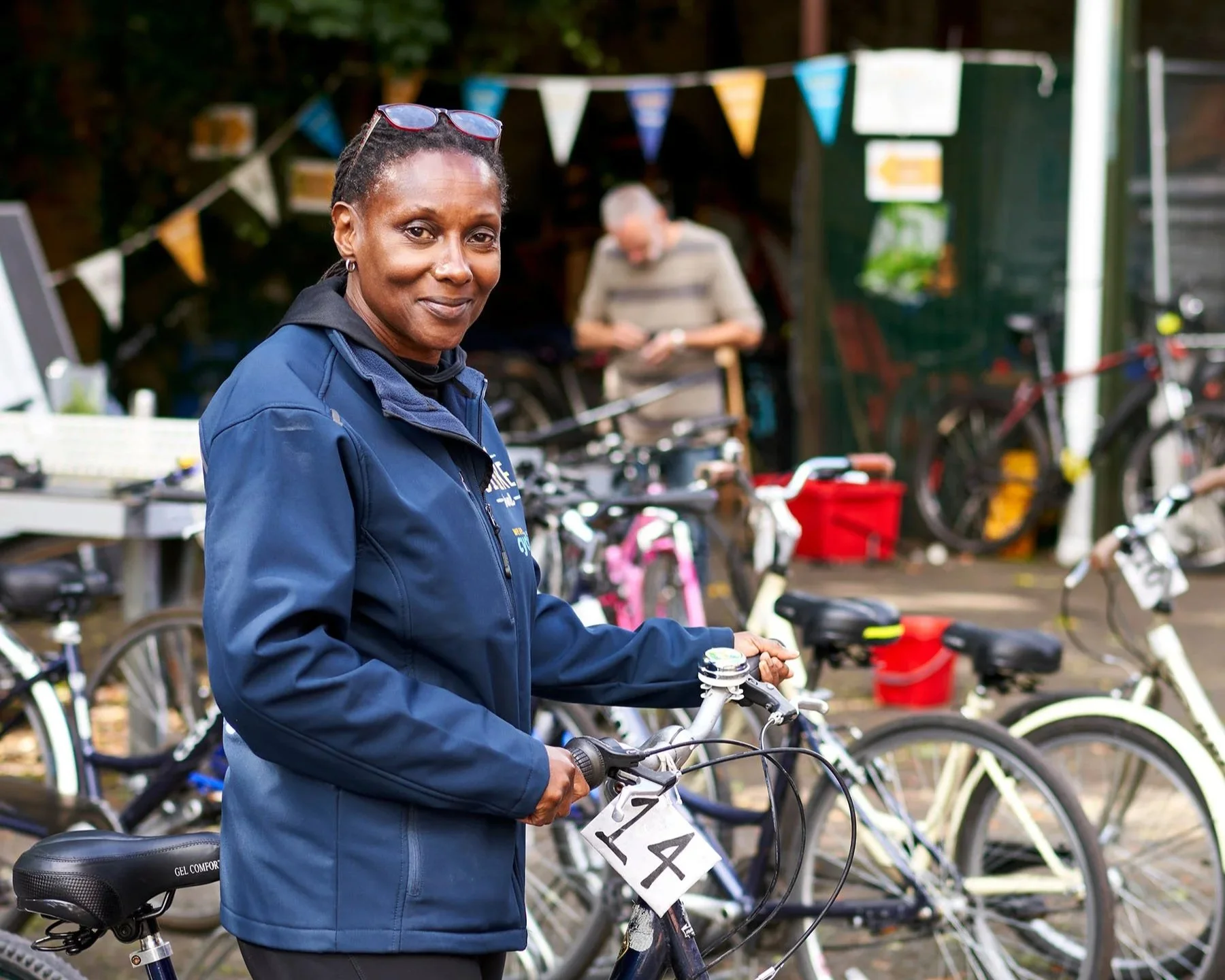 Dark skinned woman with short braided hair, sunglasses on her head, in a dark blue jacket. She is standing outside, holding bicycle handlebars, with a tag with the number 14. There are bikes, a light skinned man and bunting in the background.