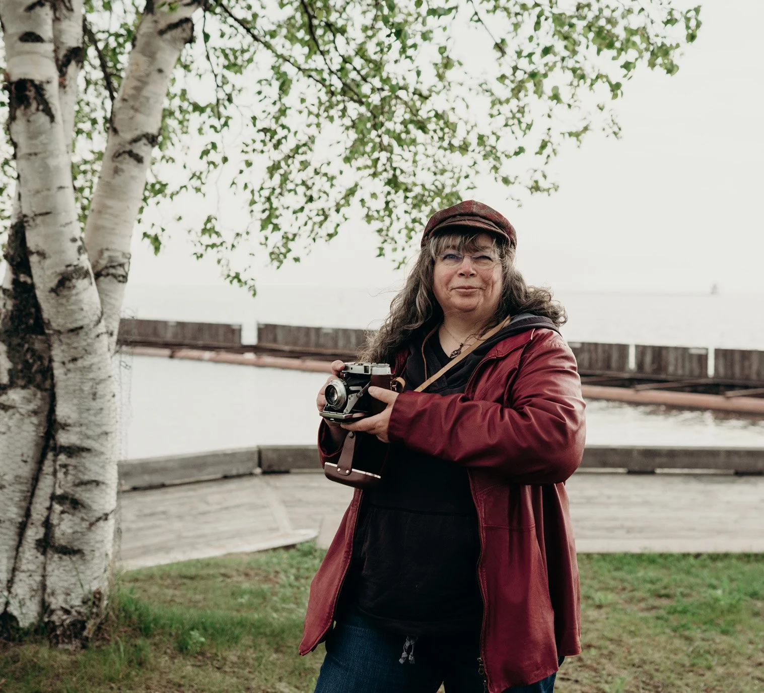 Dana wearing her cap and red leather jacket, is holding an old fashioned folding rangefinder camera while standing under some tree branches at the marina.