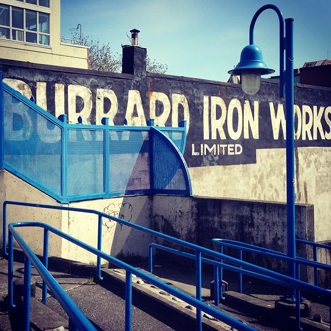 Blue railings and old fashioned light pole, in front of the Burrard Iron Words Limited sign painted on the side of a building.
