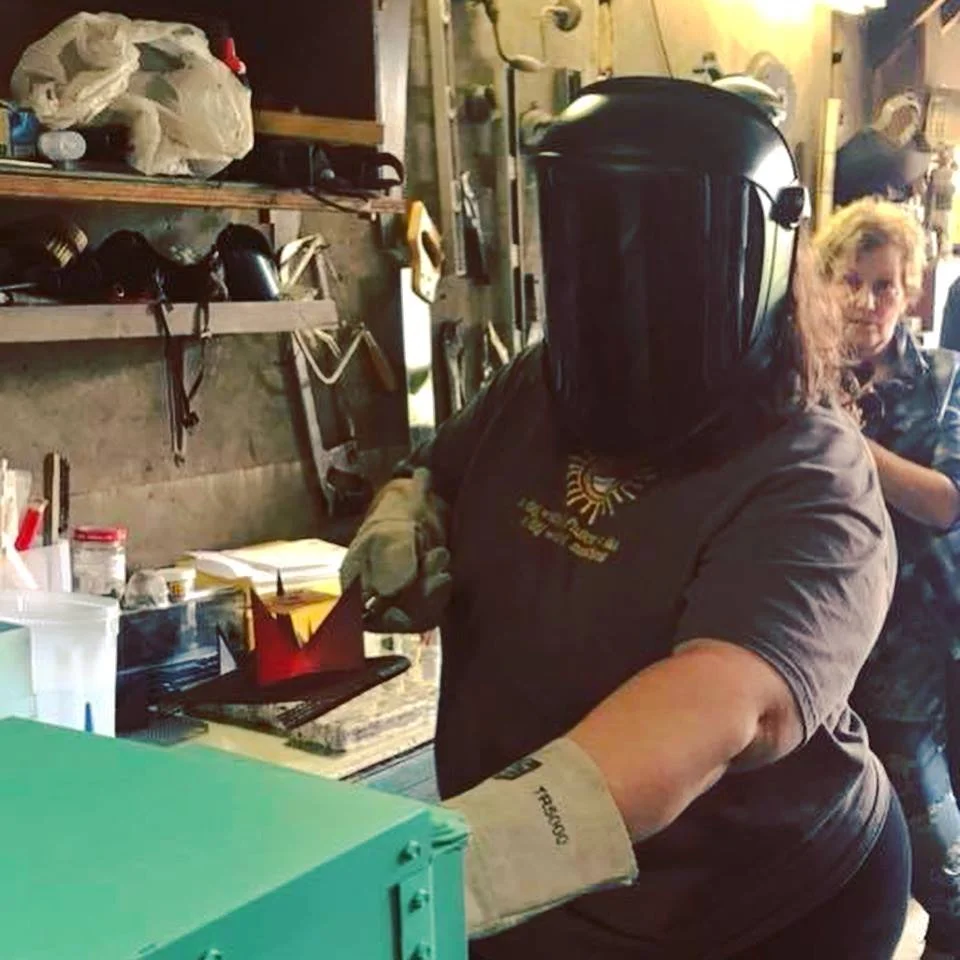 Dana wearing the same shirt and a welding mask is removing a hot steel trivet with the enamelled square on it, from a small green kiln. She is standing in a garage with tools behind her on the wall.