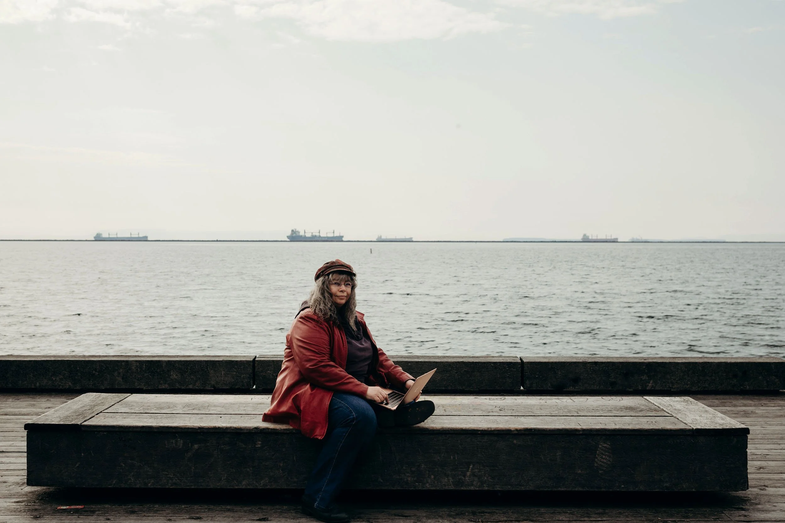 Dana Reed, a light skinned woman with long dark curly hair, wearing read leather jacket, cap and glasses, sitting on a wooden bench with Lake Superior and ships in the background.