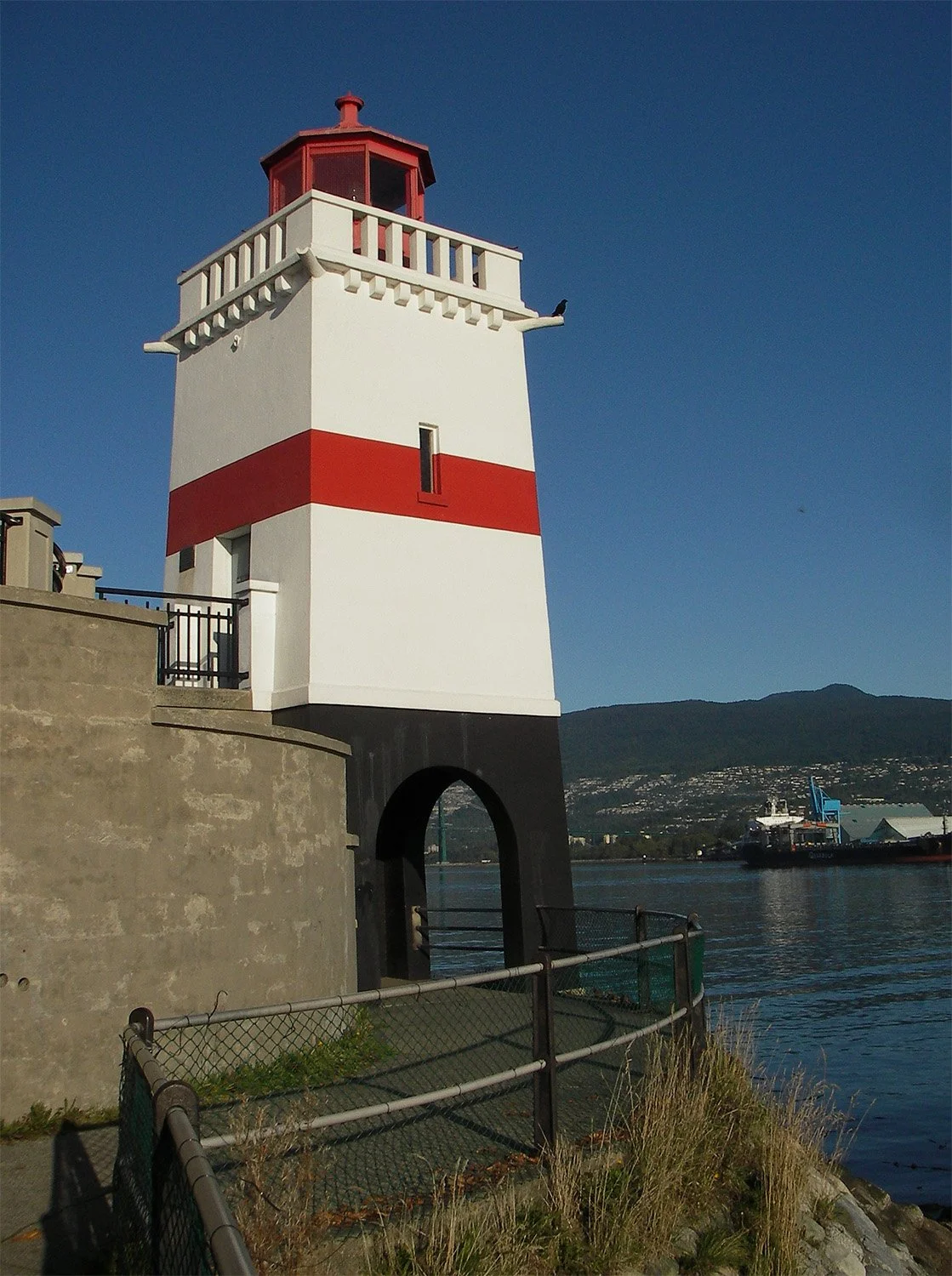 Short, square white lighthouse with red stripe on Vancouver seawall, North Vancouver and mountains in background.