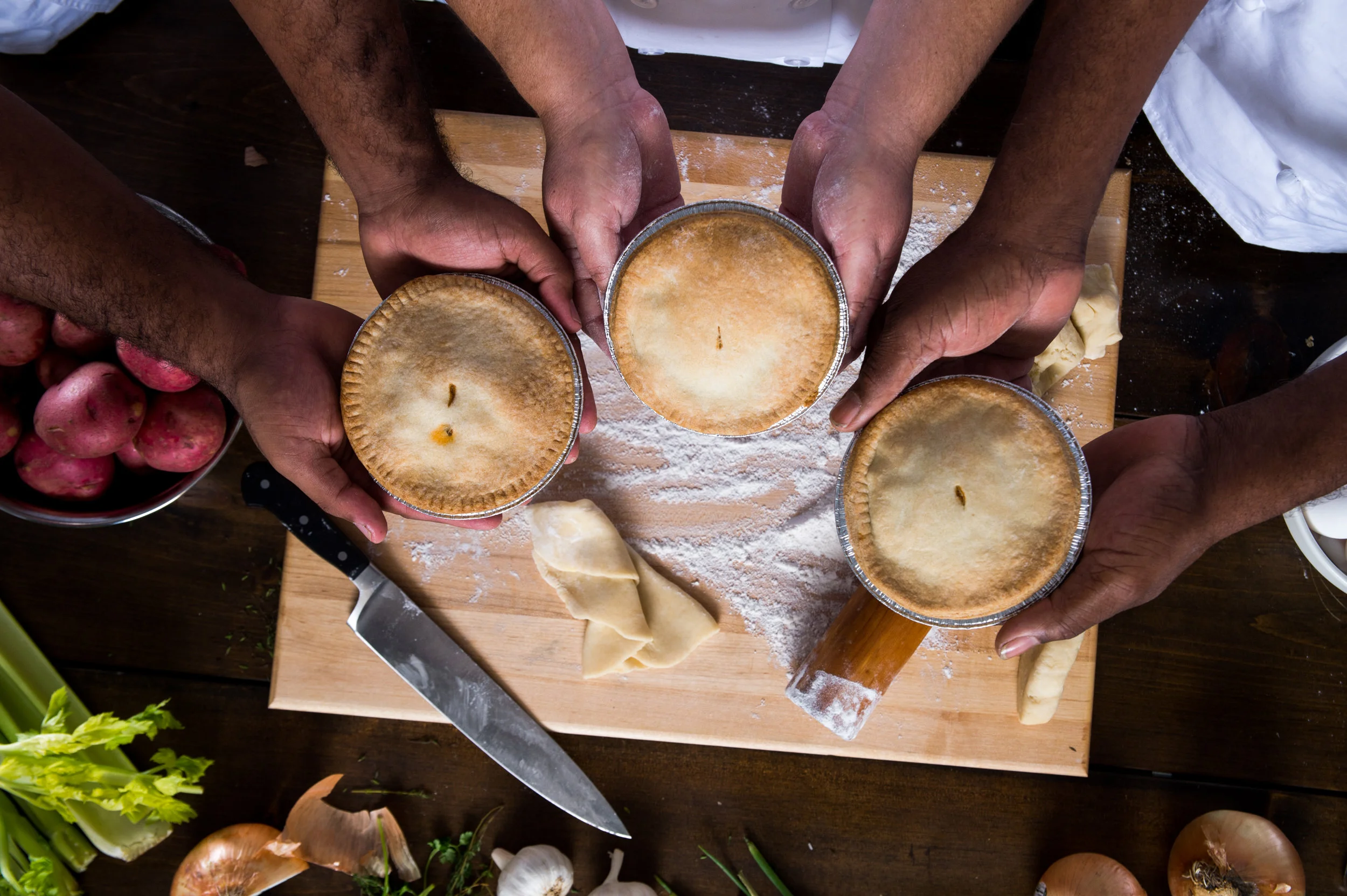 People holding drinks in foil cups, seen from above, with a wooden table covered in flour, dough, and cooking ingredients.