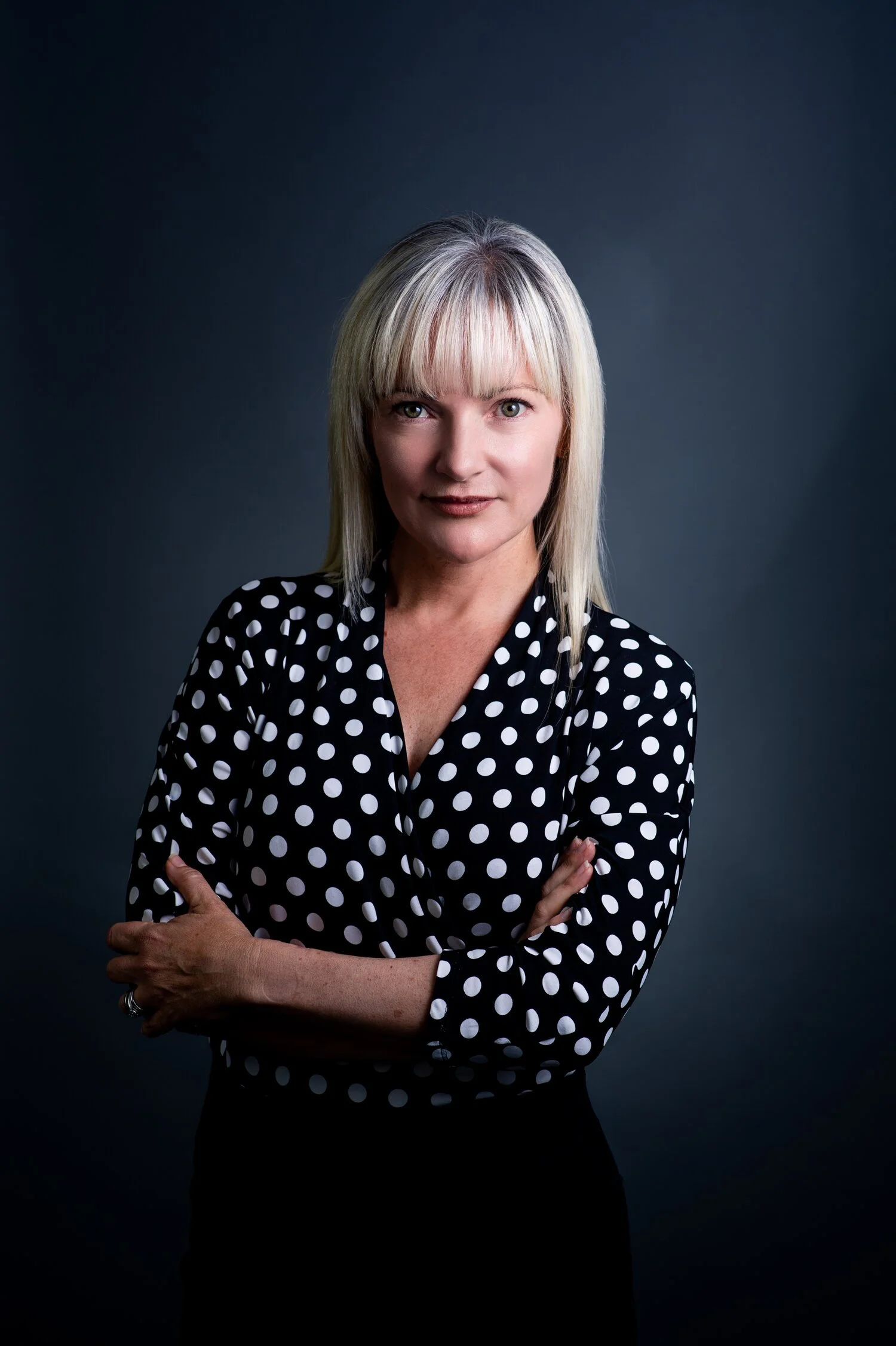 Woman with blonde hair wearing black and white polka dot shirt, arms crossed, standing against dark background.