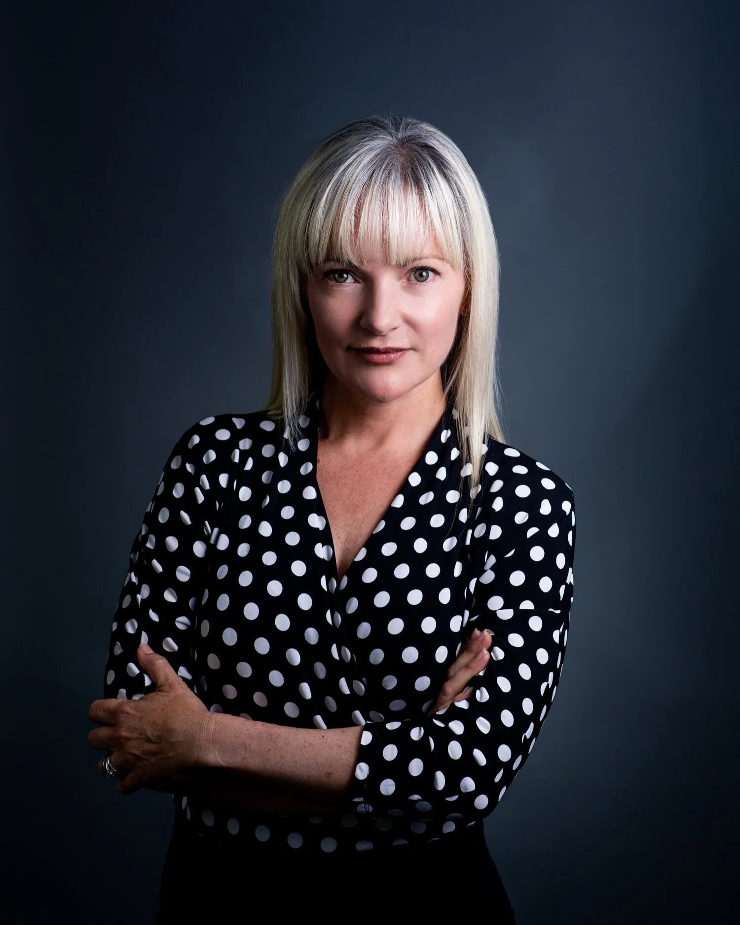 A woman with blonde hair, wearing a black blouse with white polka dots, standing with arms crossed against a dark background.