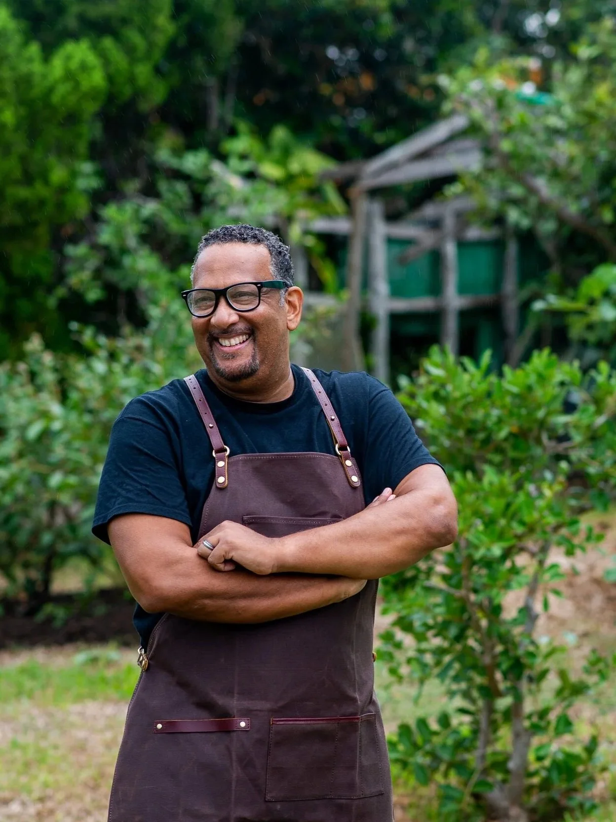 A man smiling and standing outdoors in a garden, wearing glasses, a black t-shirt, and a brown apron with his arms crossed, surrounded by green bushes and trees.