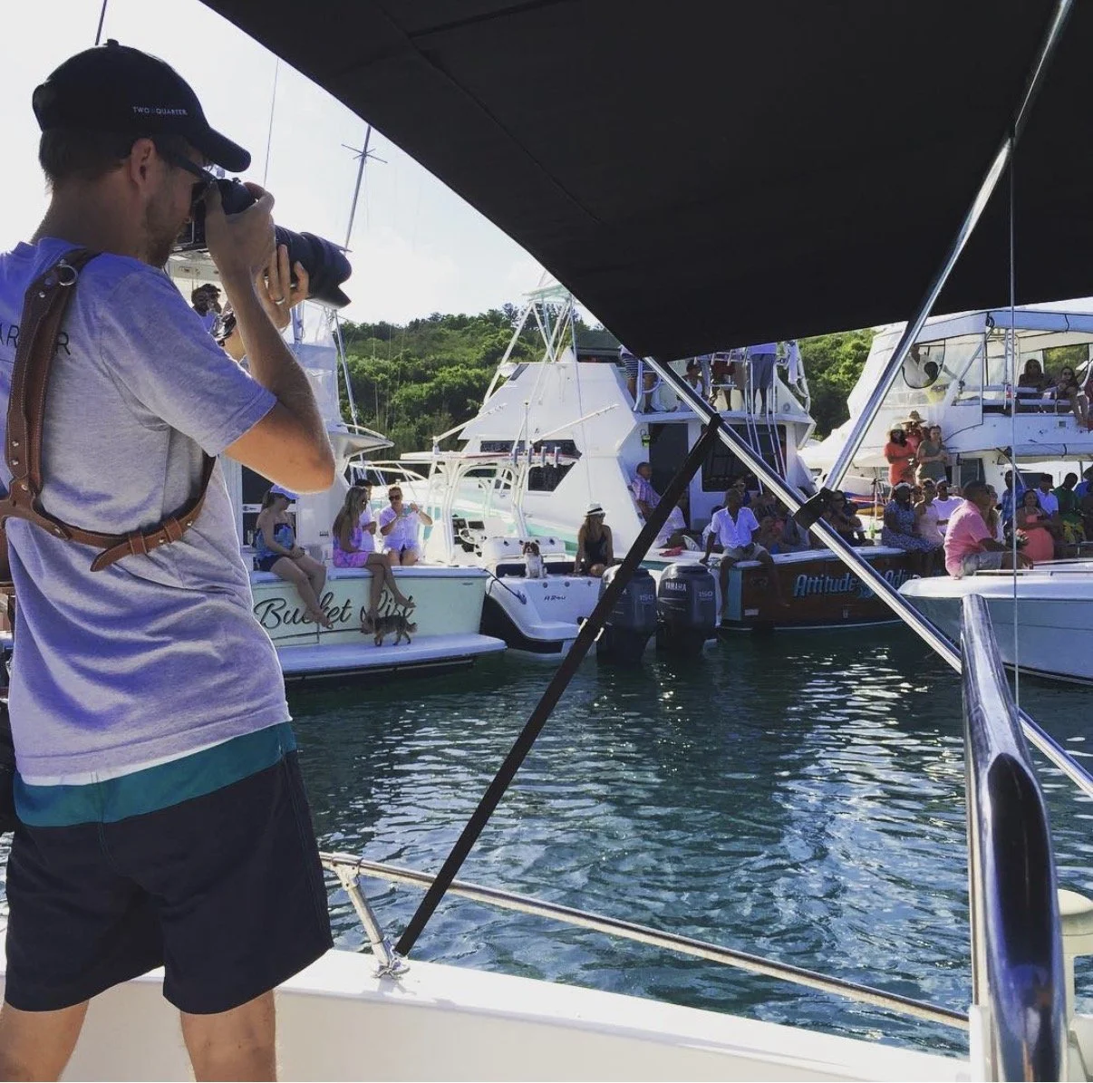 A man with a camera aboard a boat, taking a photo of a busy marina with multiple boats and people enjoying a day on the water, with green hillside in the background.