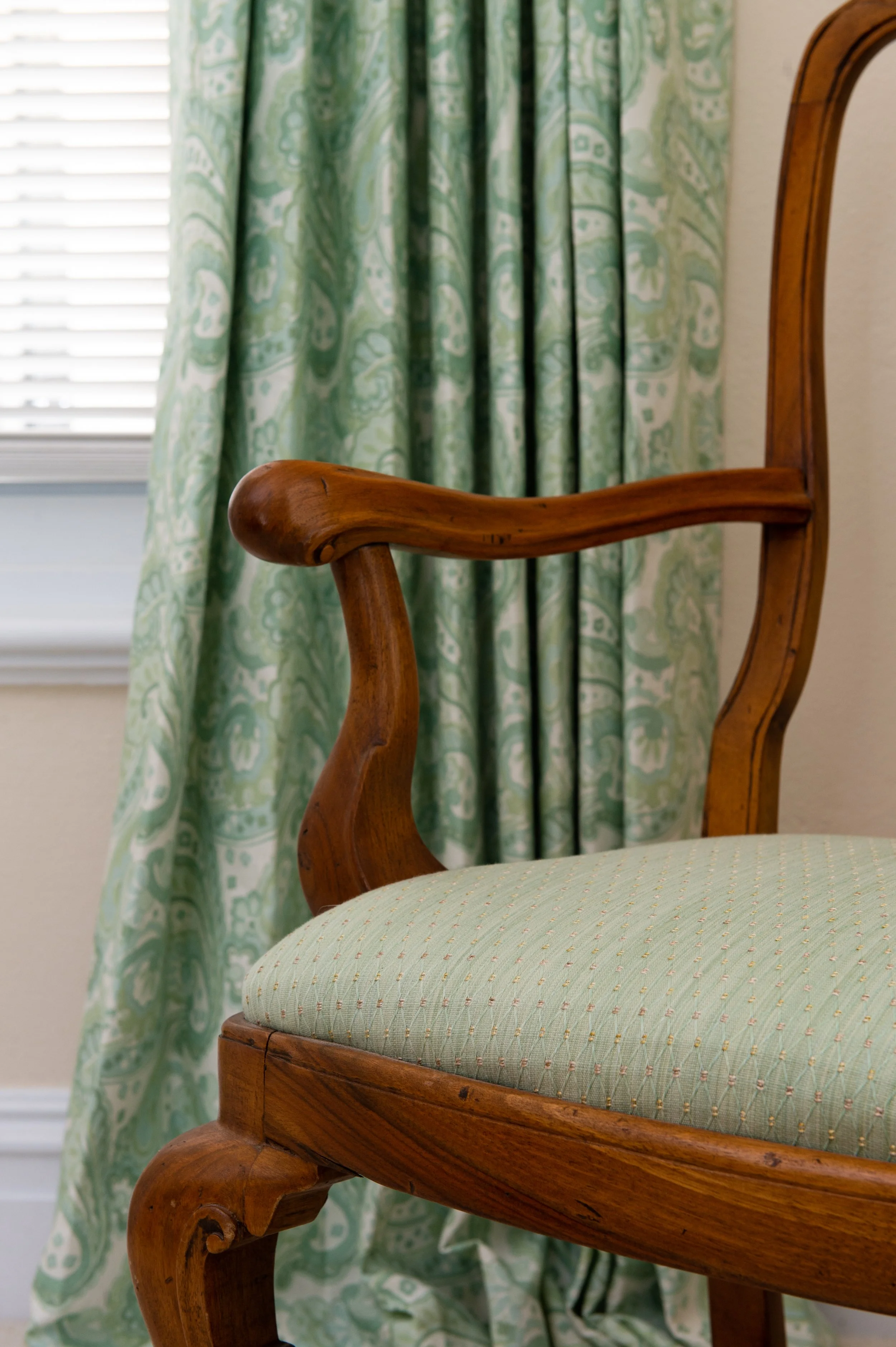 Close-up of a vintage wooden armchair with a light green, dotted upholstered seat, positioned near a window with a curtain featuring a green and white pattern.