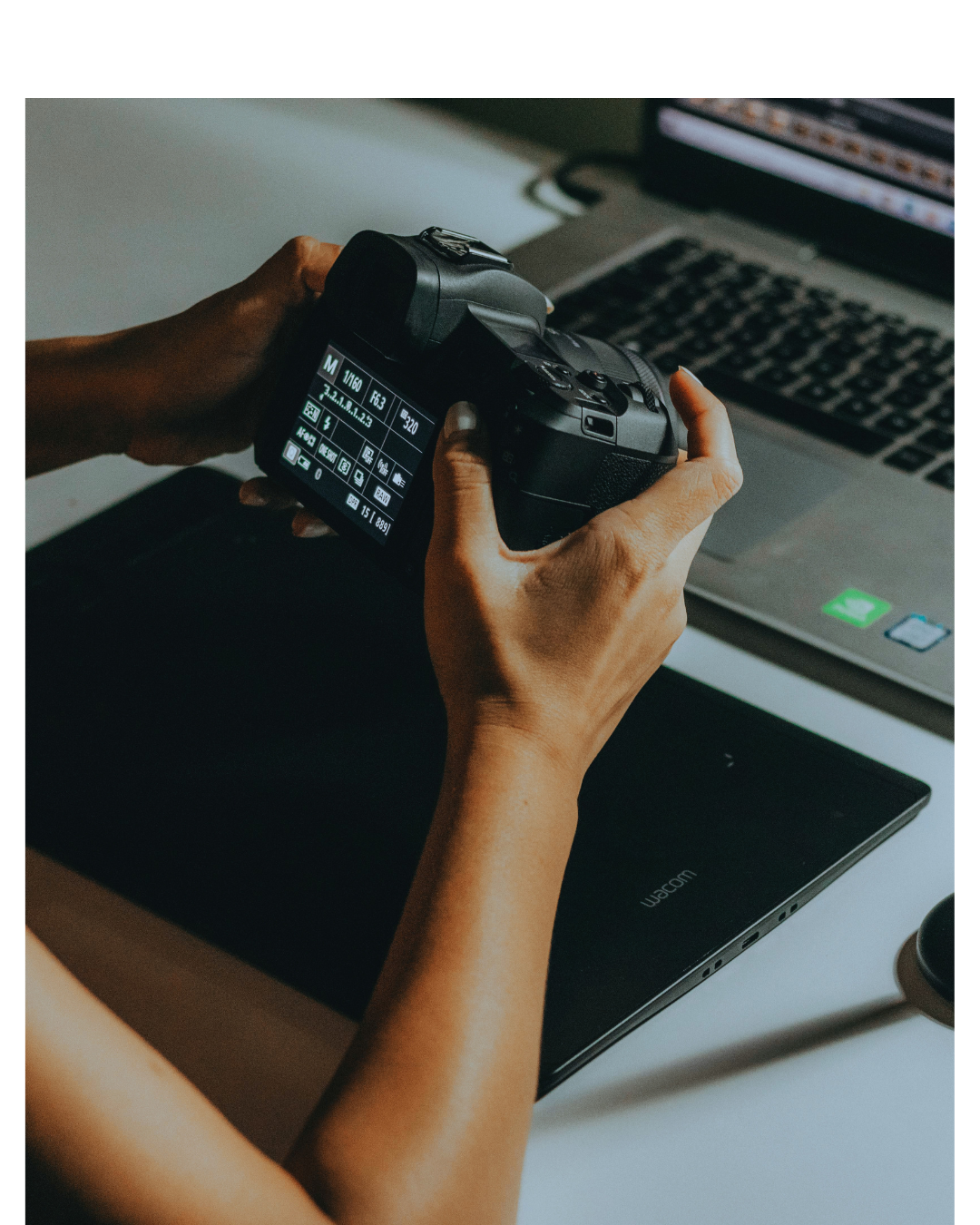 A person holding a digital camera over a closed Wacom tablet, with a laptop in the background.