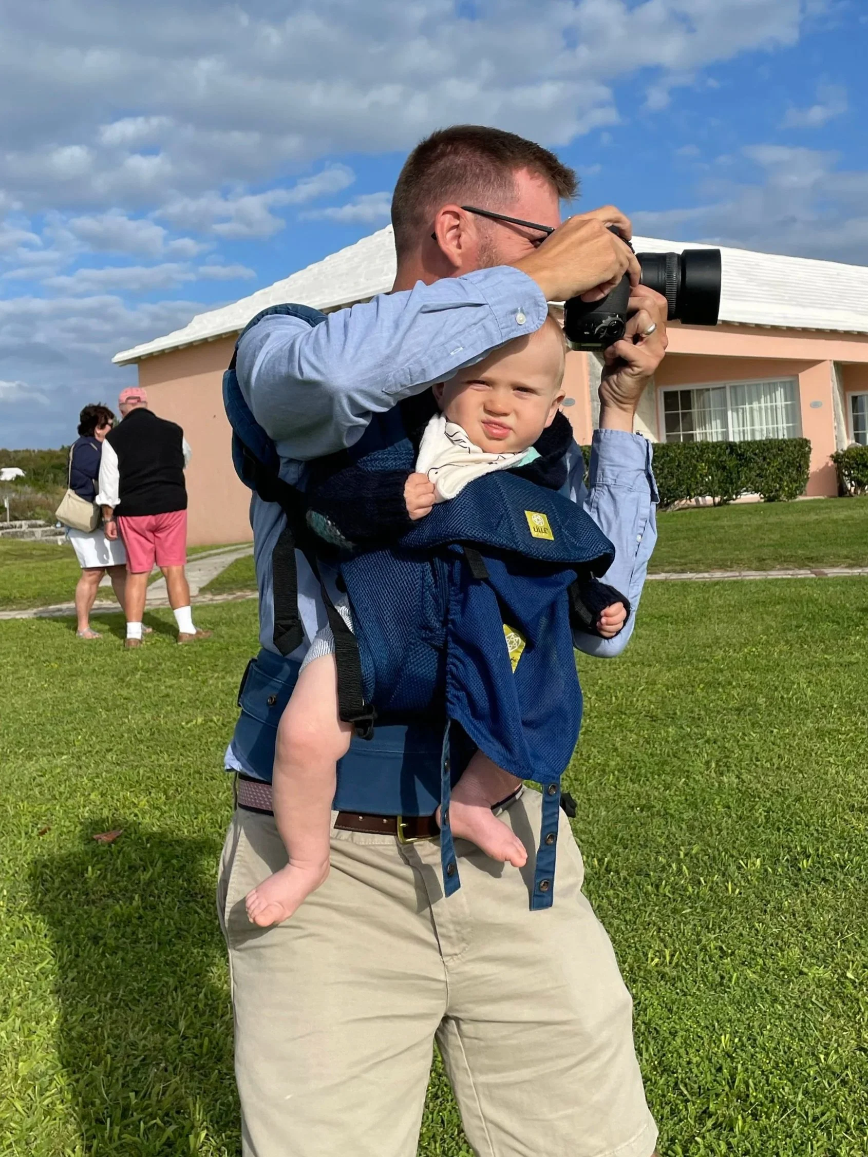 Man carrying a baby in a blue backpack slung over his shoulders, using a camera to take a photo outdoors on a grassy lawn near a pink building with a white roof, with a cloudy sky in the background.