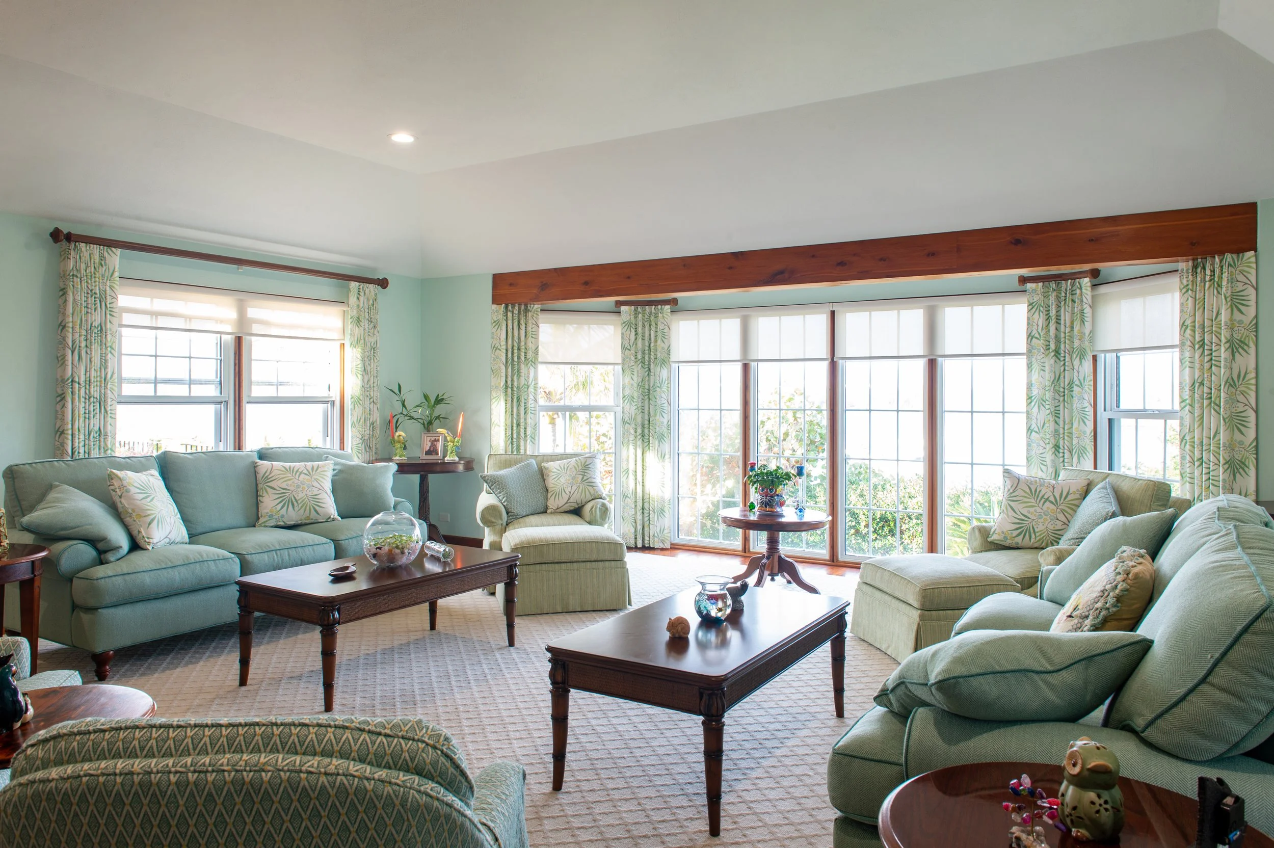 Bright living room with large windows, light green furniture, patterned curtains, and wooden coffee tables.