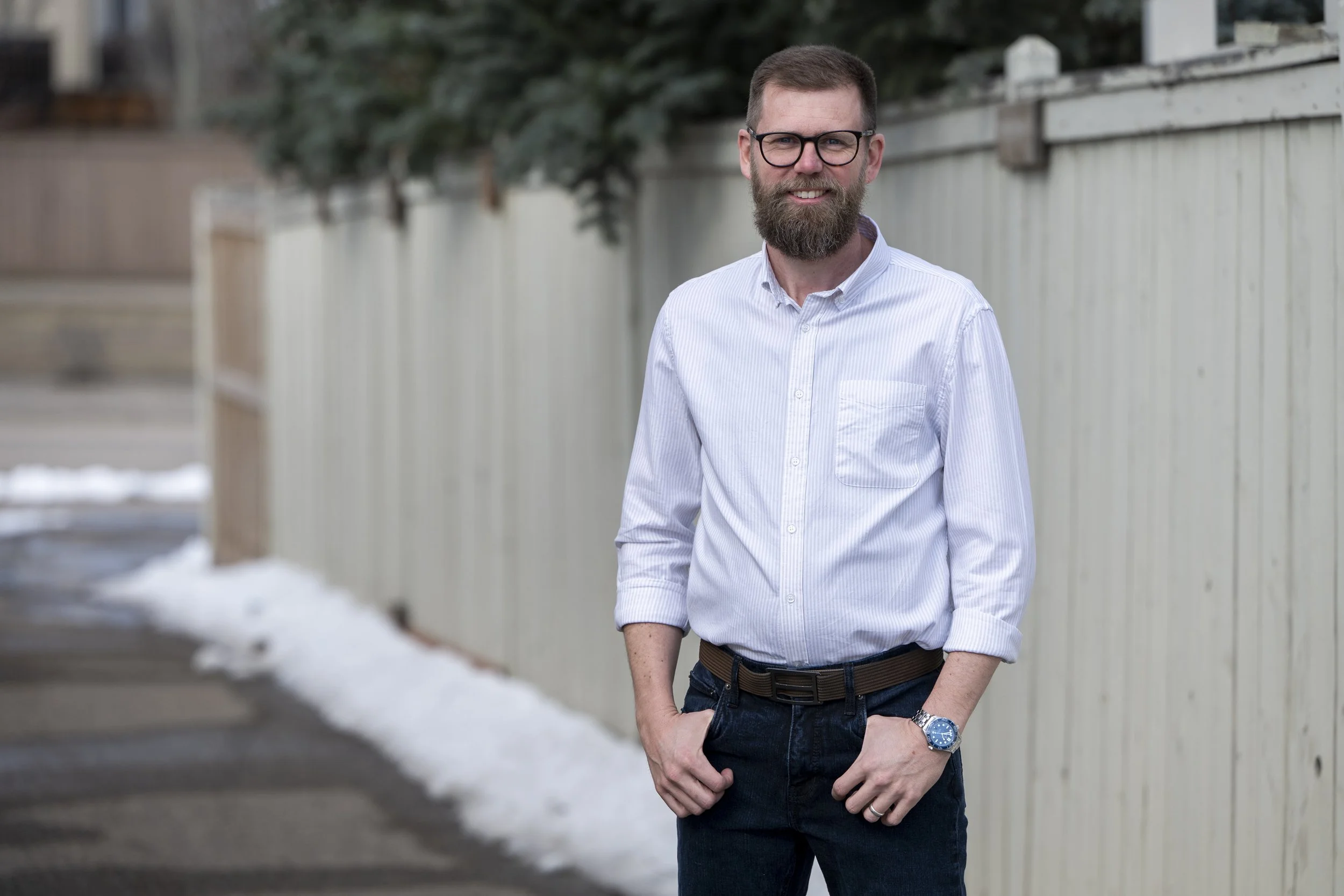 A man with glasses and a beard smiling outdoors, wearing a white button-down shirt and dark pants, standing in front of a white fence with snow on the ground.