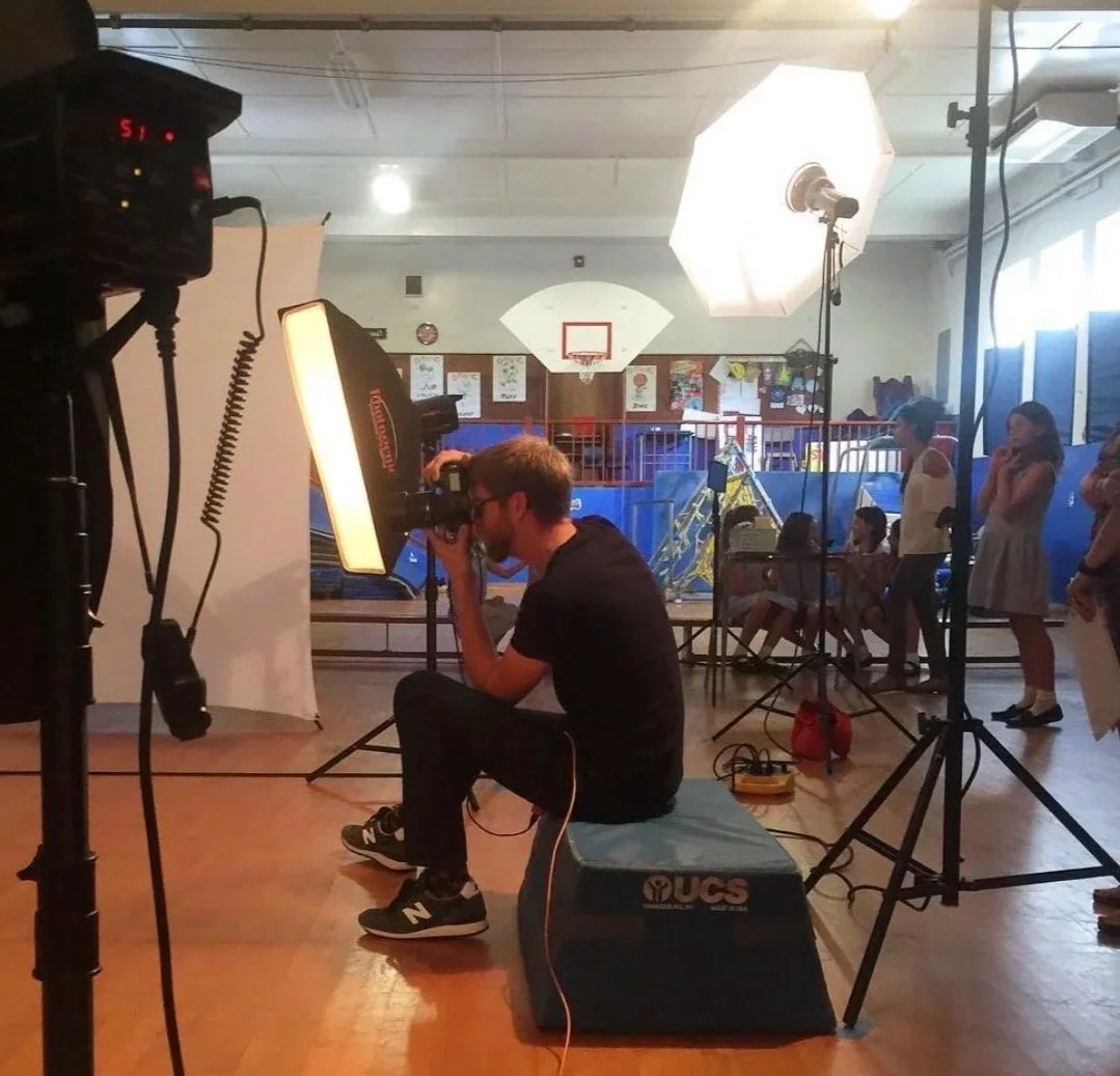 Photographer taking a picture of children in a gymnasium with basketball hoops, using professional lighting equipment and a backdrop.