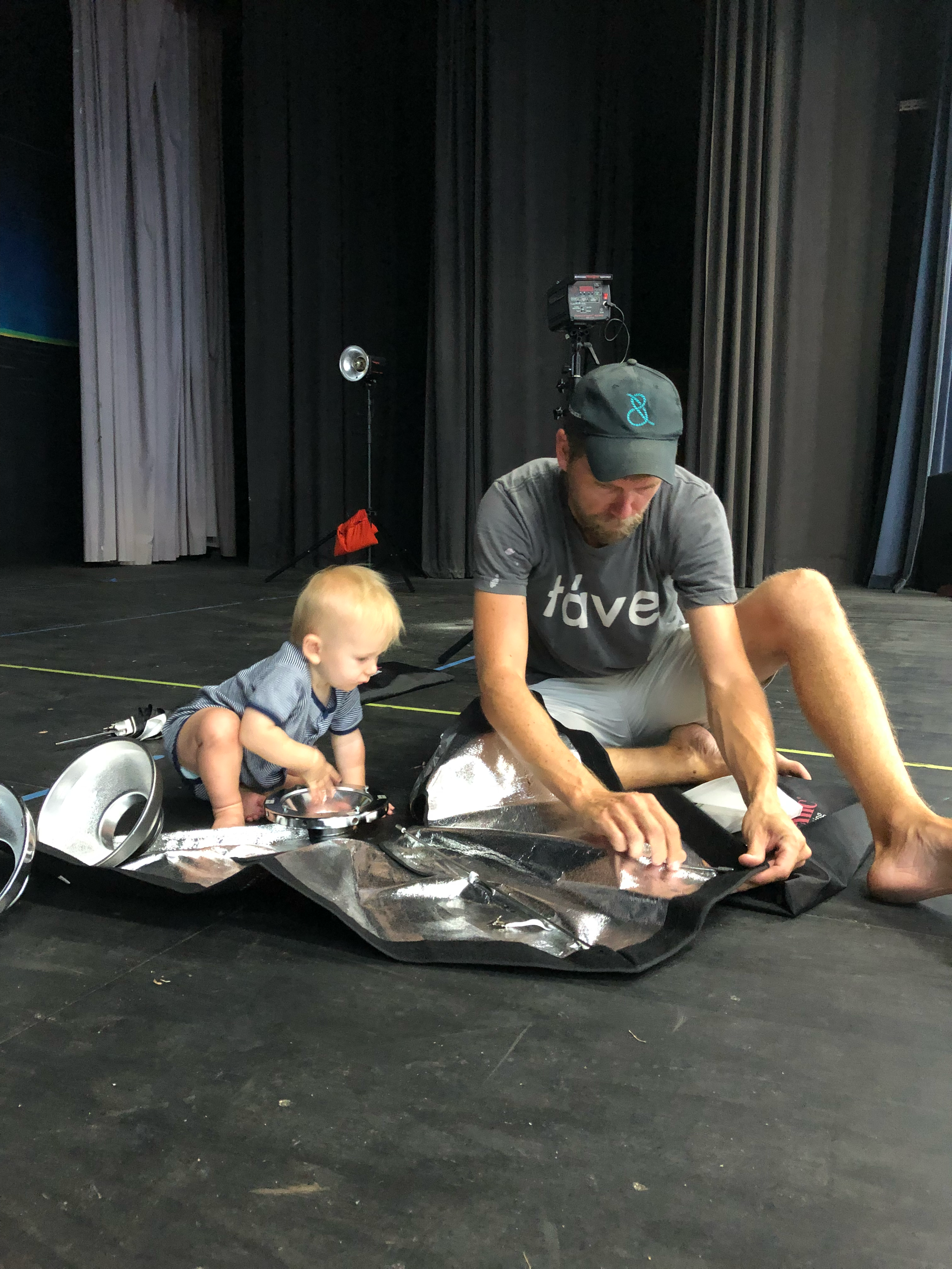 Man and young child sitting on stage floor with silver metallic foil material and equipment around them, dark curtains in background.