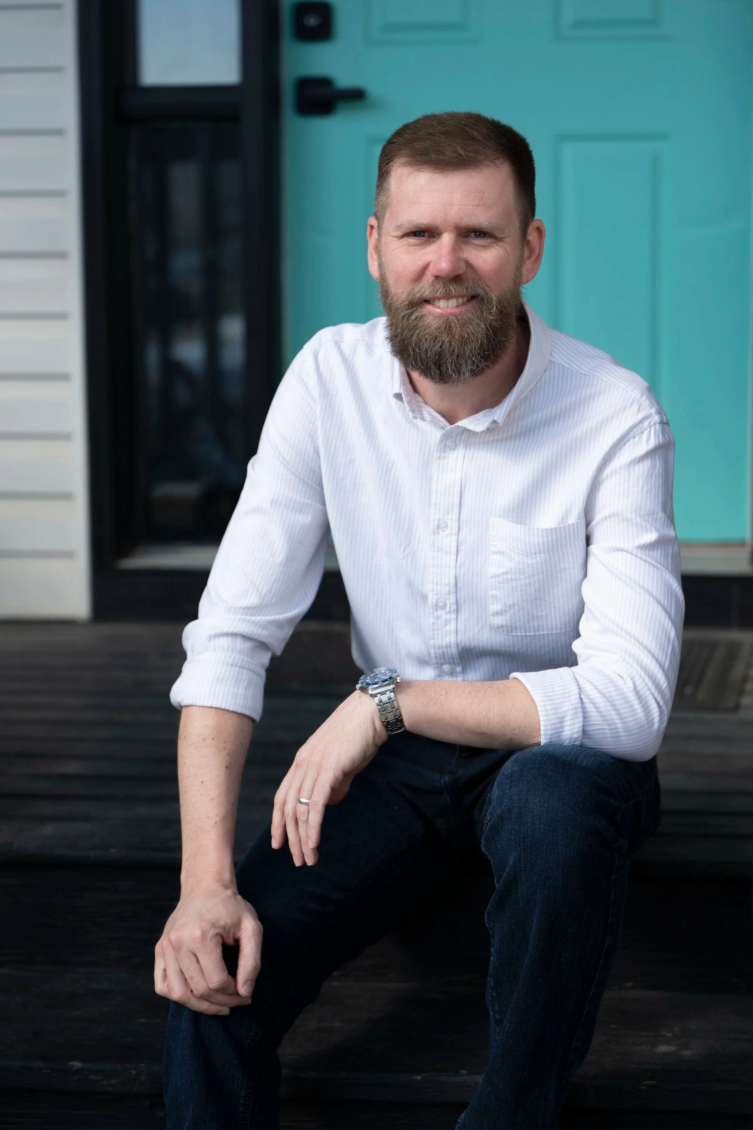 A man with a beard and short hair, wearing a white button-up shirt and dark jeans, is sitting on a wooden deck in front of a teal door, smiling at the camera.